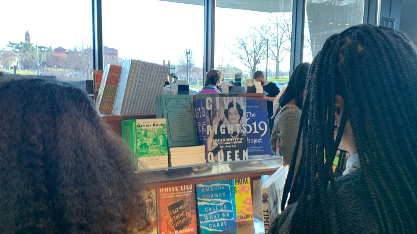 View of people shopping for books inside a bookstore with large windows showing an outdoor scene with trees and buildings.