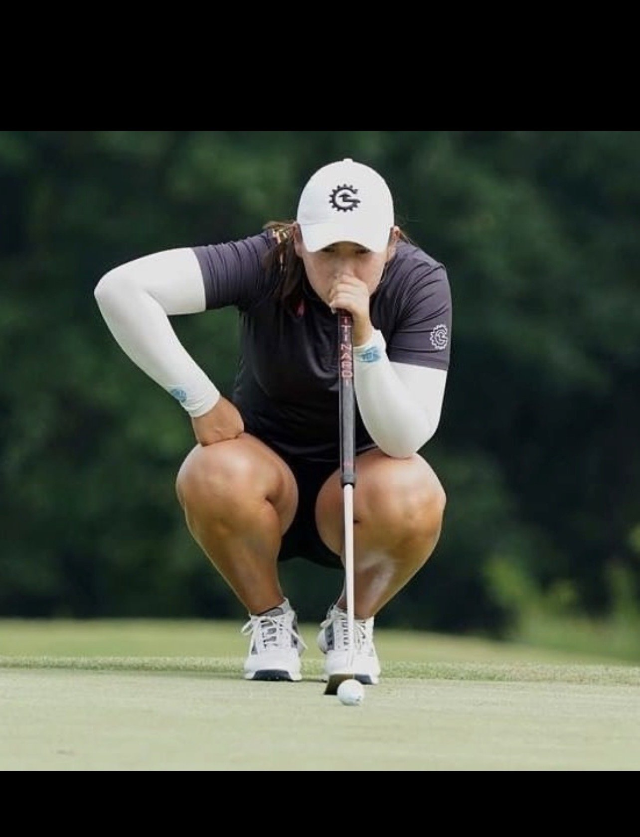 A woman crouching on a golf course, lining up her shot with a golf tee and golf ball, surrounded by a green background.