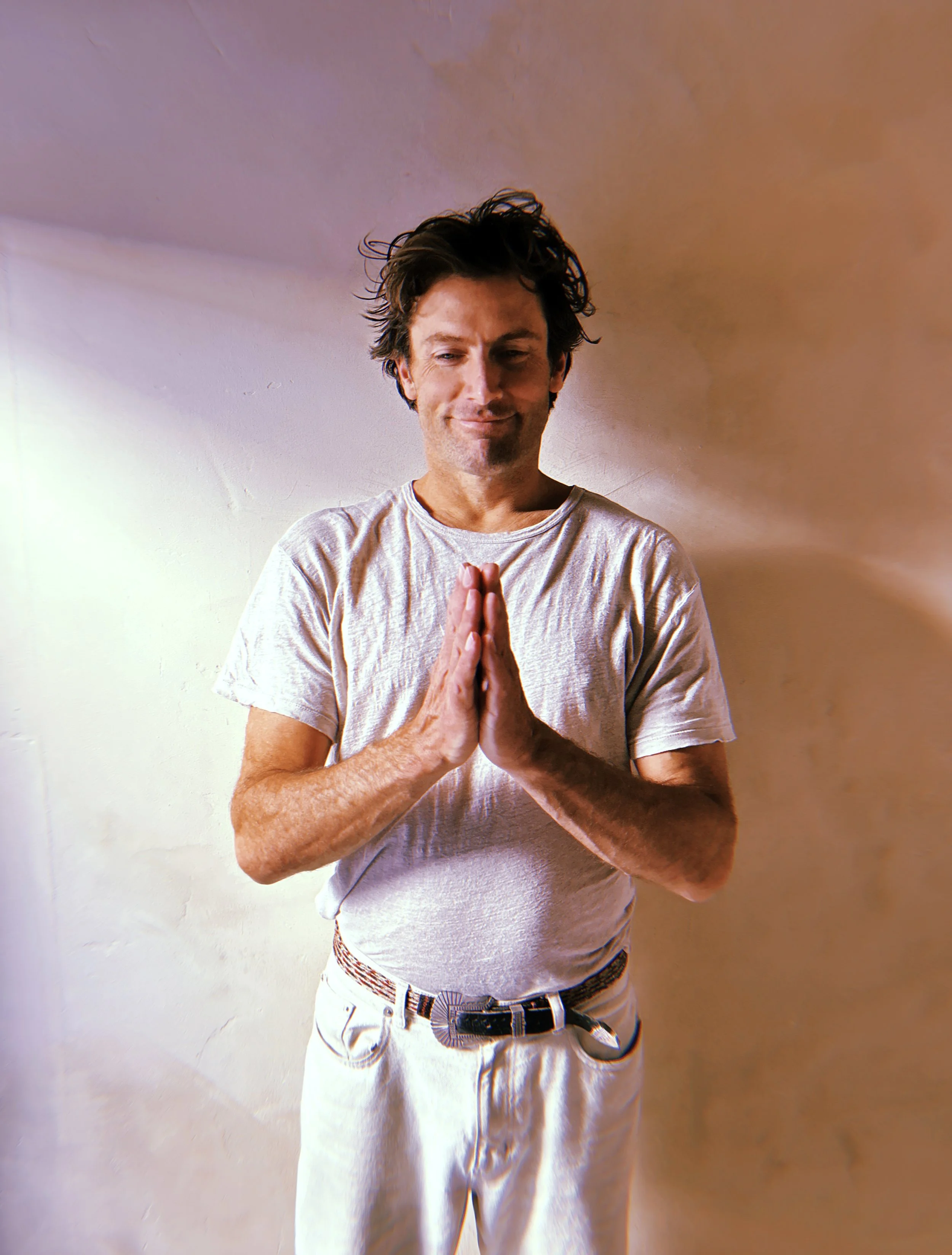 KC Deane with dark curly hair standing indoors against a plain wall, holding his hands together in a prayer position, with a serene expression.