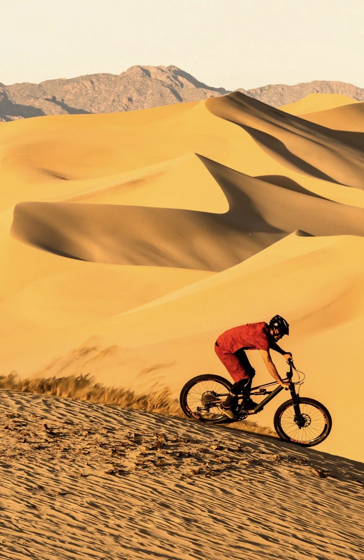 KC Deane  rides a bicycle at the sandy dumont dunes recreation area with mountains in the background during sunset.