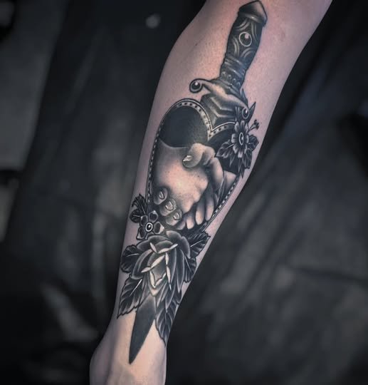 Tattoo of a hand gripping a dagger, surrounded by flowers, on a person's forearm.