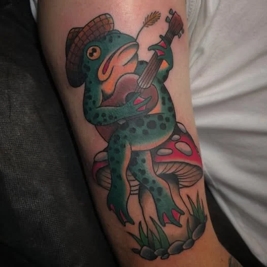 Tattoo of a frog sitting on a mushroom, wearing a cowboy hat, playing a guitar, with a grass area and rocks at the base.