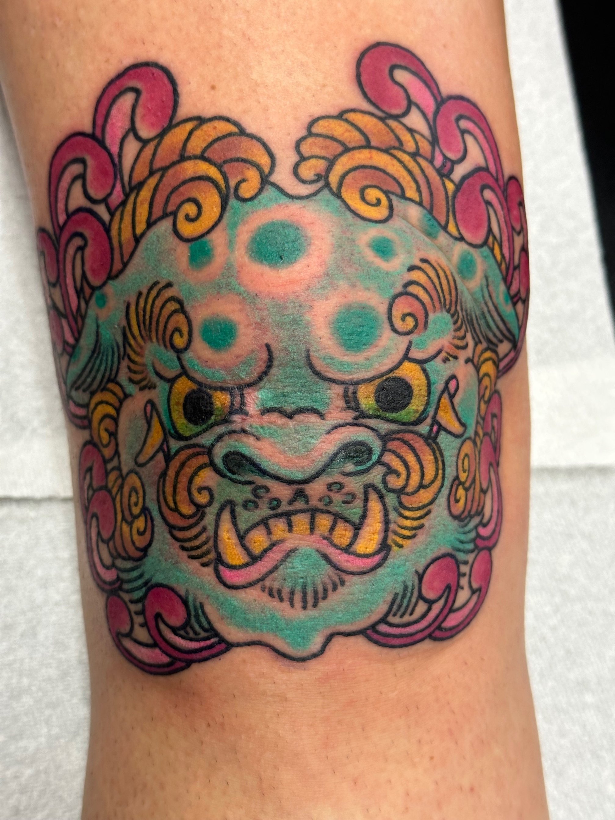 Colorful tattoo of a fierce mythical creature's face with swirling patterns, prominent teeth, and bright colors.