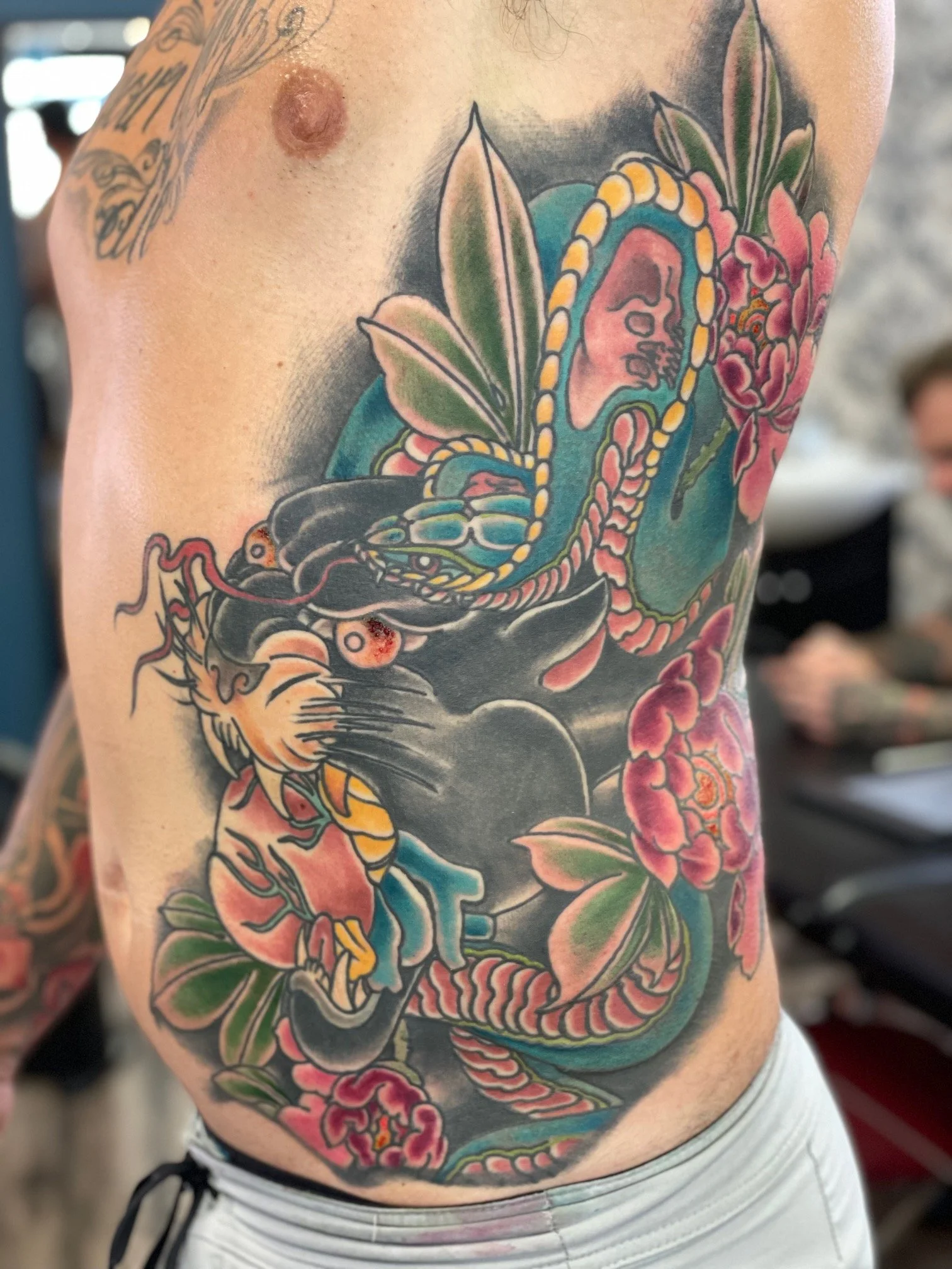 Colorful tattoo on person's side featuring a tiger, a snake, flowers, leaves, and a face inside the snake's body.