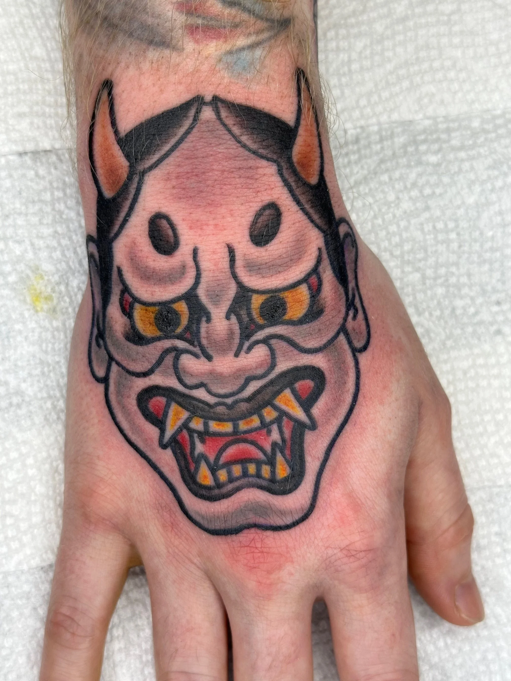 A freshly inked tattoo of a Japanese Hannya mask on a person's hand, featuring fierce facial features, sharp teeth, and a demonic expression, with a background of white textured paper or paper towel.