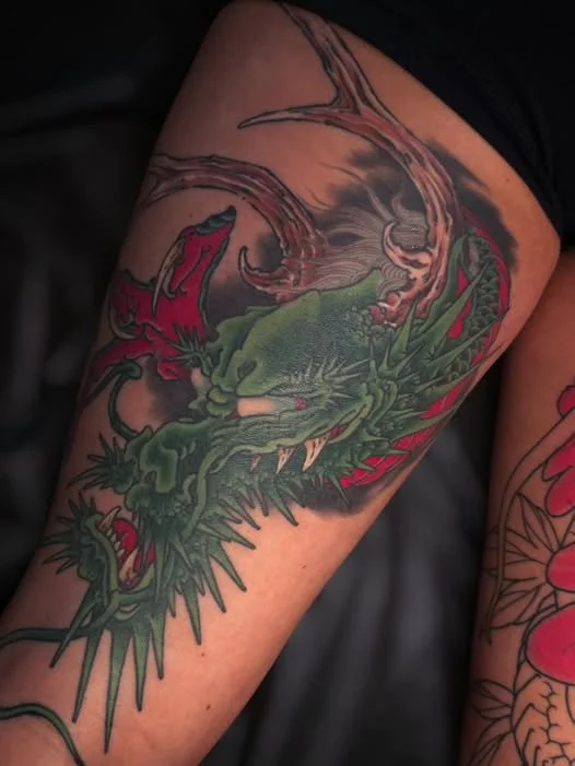 A tattoo of a green dragon with red accents on a person's thigh, surrounded by smoke or clouds.