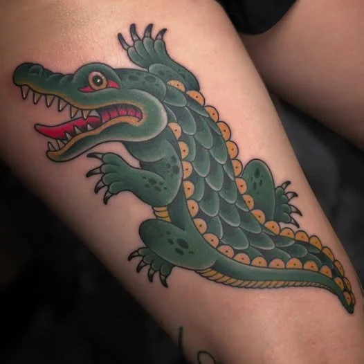 Tattoo of a cartoon-style alligator with an open mouth, showing teeth, and colorful details on skin and tail.