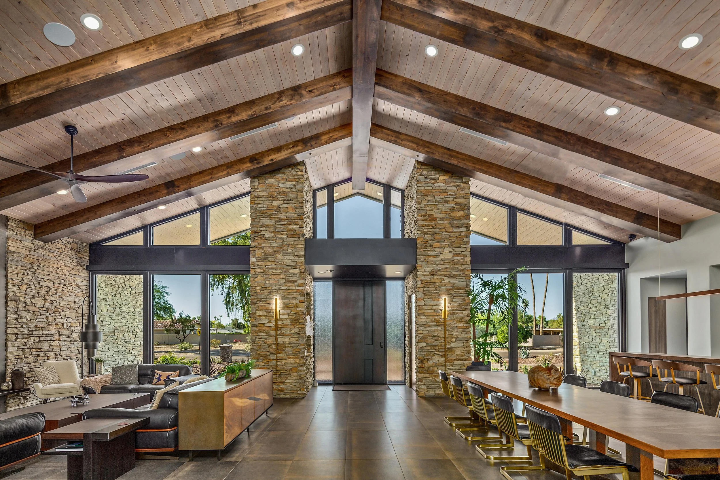 Interior of a modern living room with a high vaulted ceiling, exposed wood beams, stone walls, large windows, and contemporary furniture.