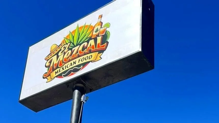 A sign for El Mezcal Mexican Food featuring colorful graphics of agave leaves, a bottle, and a Mexican sombrero against a clear blue sky.