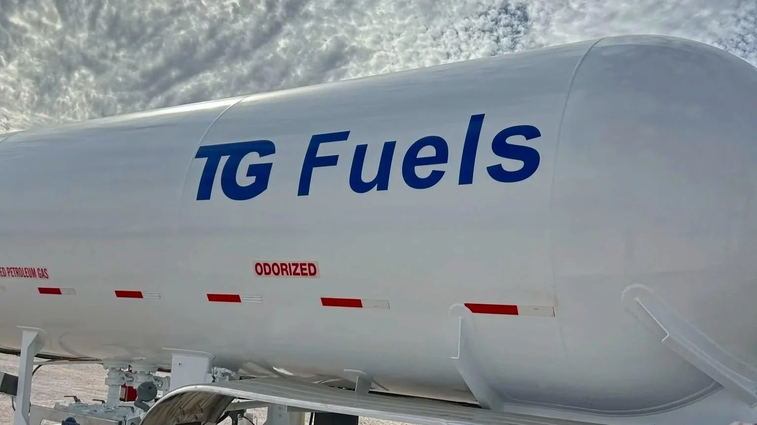 Close-up of a white tank labeled 'TG Fuels' with red warning labels, on a snowy landscape with a cloudy sky in the background.