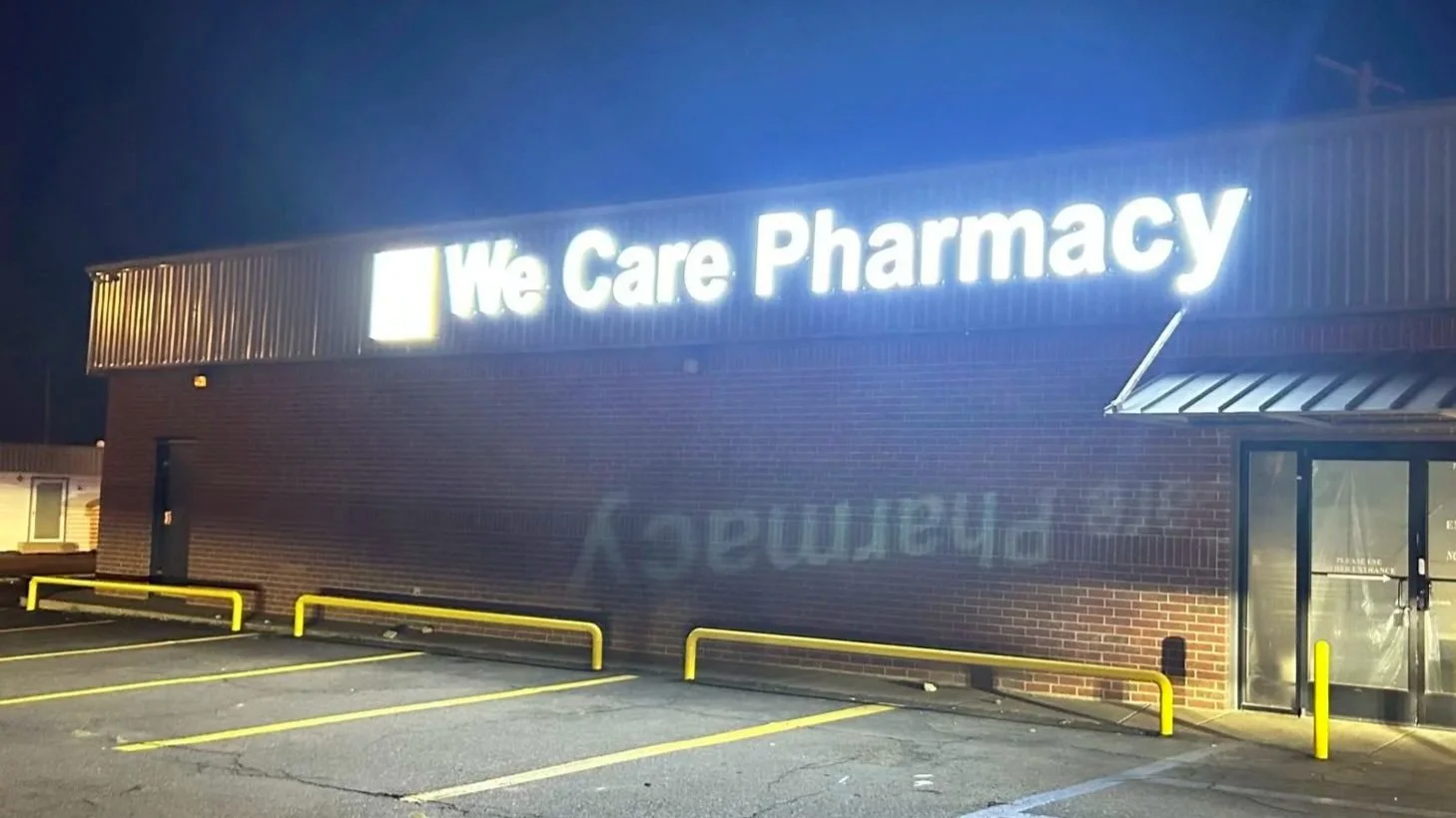 The exterior of a pharmacy store named 'We Care Pharmacy' at night with illuminated sign, brick walls, yellow parking lot barriers, and an entrance door.