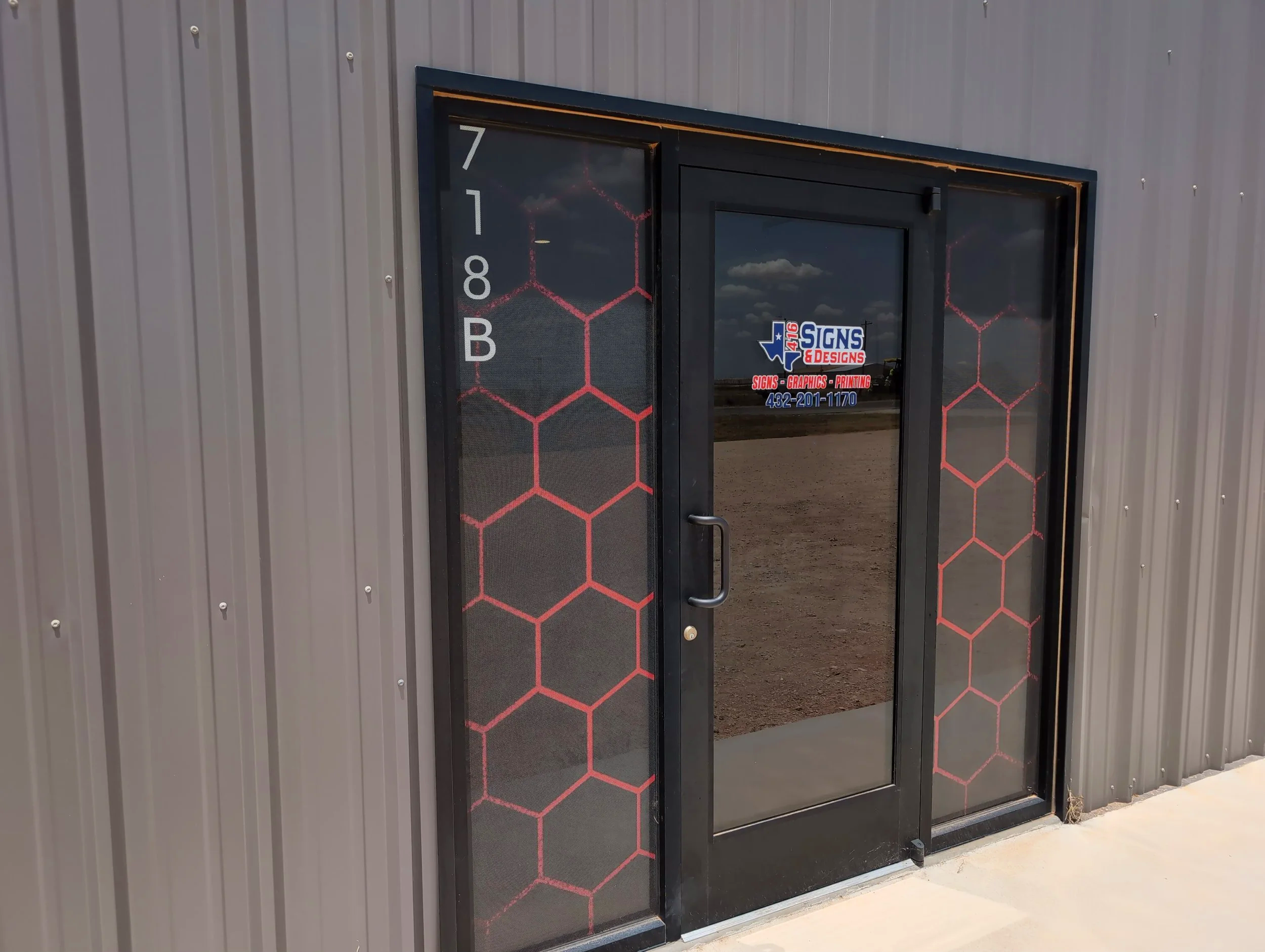 Exterior view of a building with a glass door and side panels, featuring signage for Texas Signs & Designs, a business specializing in signs, graphics, and printing. The side panels have a red honeycomb pattern, and the building has gray vertical siding.