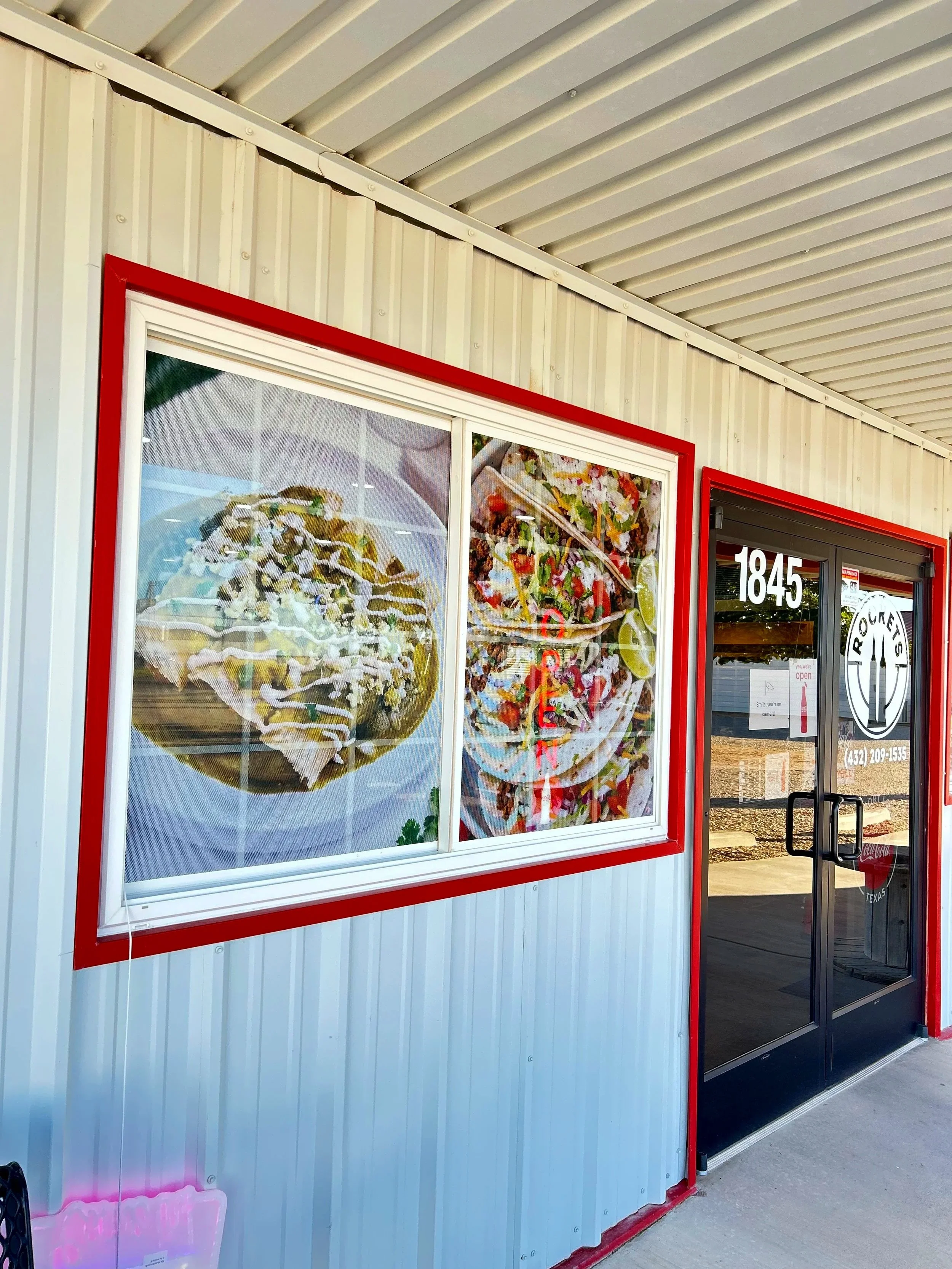 Outdoor view of a restaurant with two large menu posters displaying pictures of food items, including tacos and bowls, in black frames with red borders. The restaurant's entrance has a glass door with the street number 1845 and a logo that reads 'Rocky's'. The building has cream-colored vertical siding and a metal awning above the entrance.