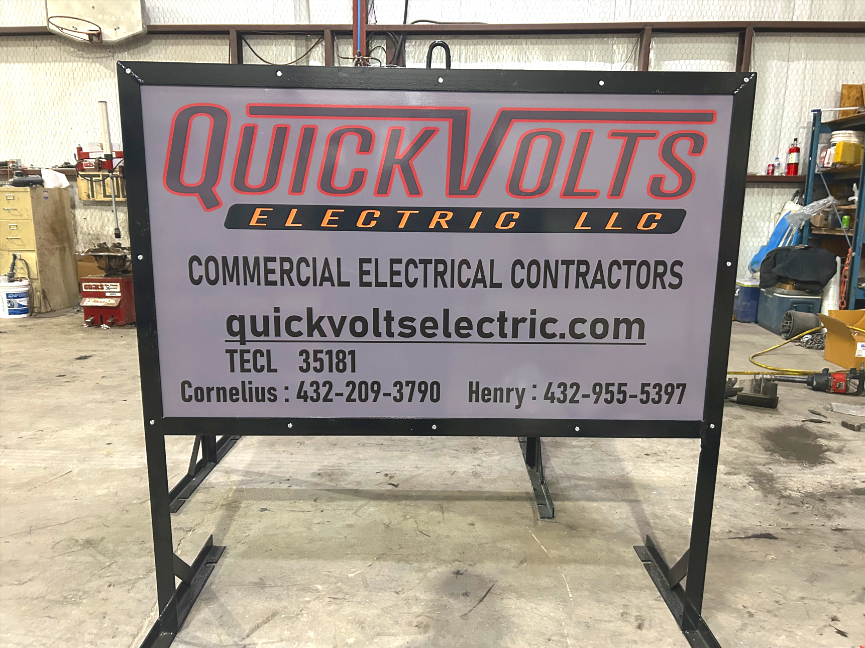 A large black-framed outdoor sign for Quick Volt Electric LLC in a workshop or warehouse setting. The sign displays the company name in large red and black letters along with the description 'Commercial Electrical Contractors,' the website quickvoltsellectric.com, and contact phone numbers for Cornelius and Henry.