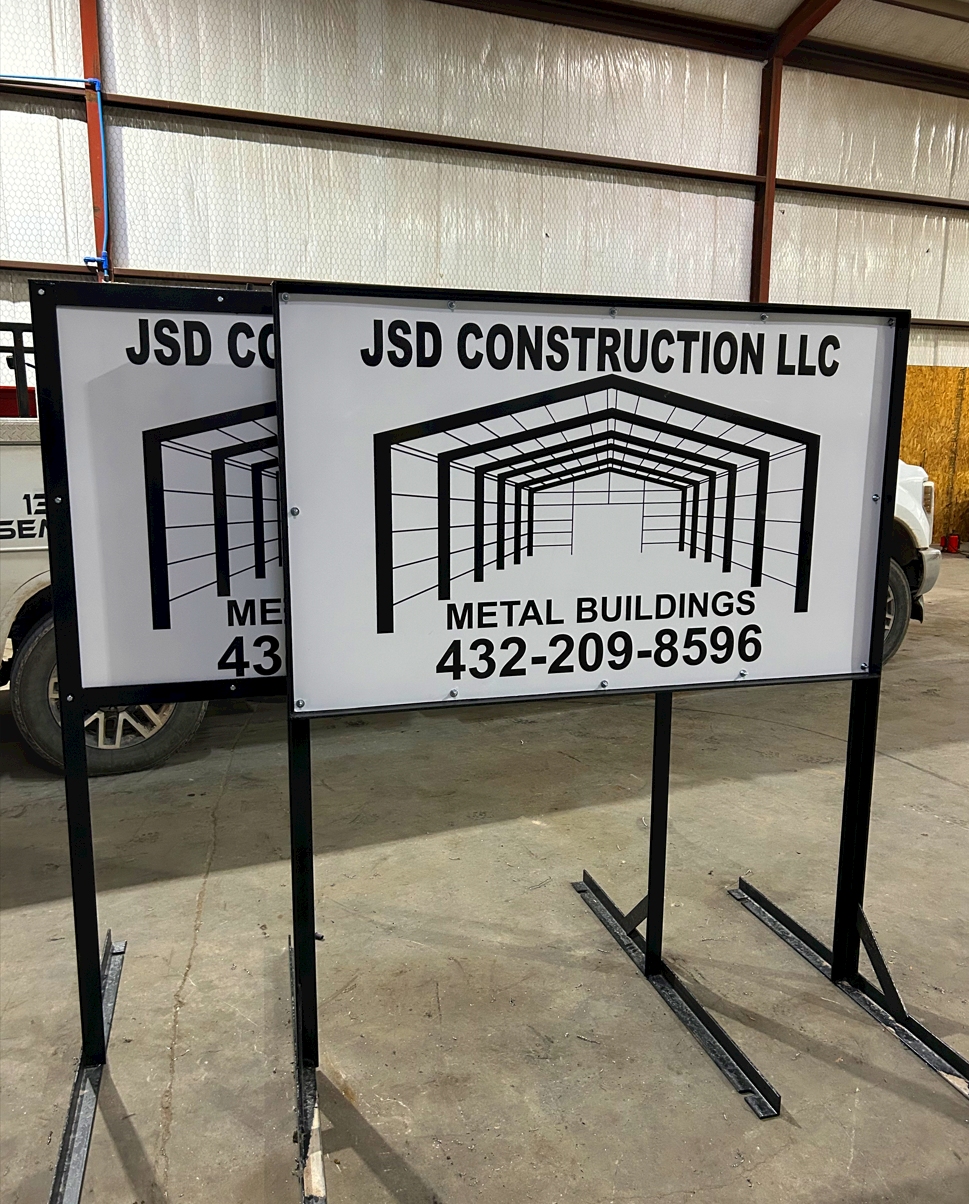 Two outdoor signs for JSD Construction LLC with a graphic of a metal building framework and contact information, placed on a concrete floor inside a warehouse or garage.