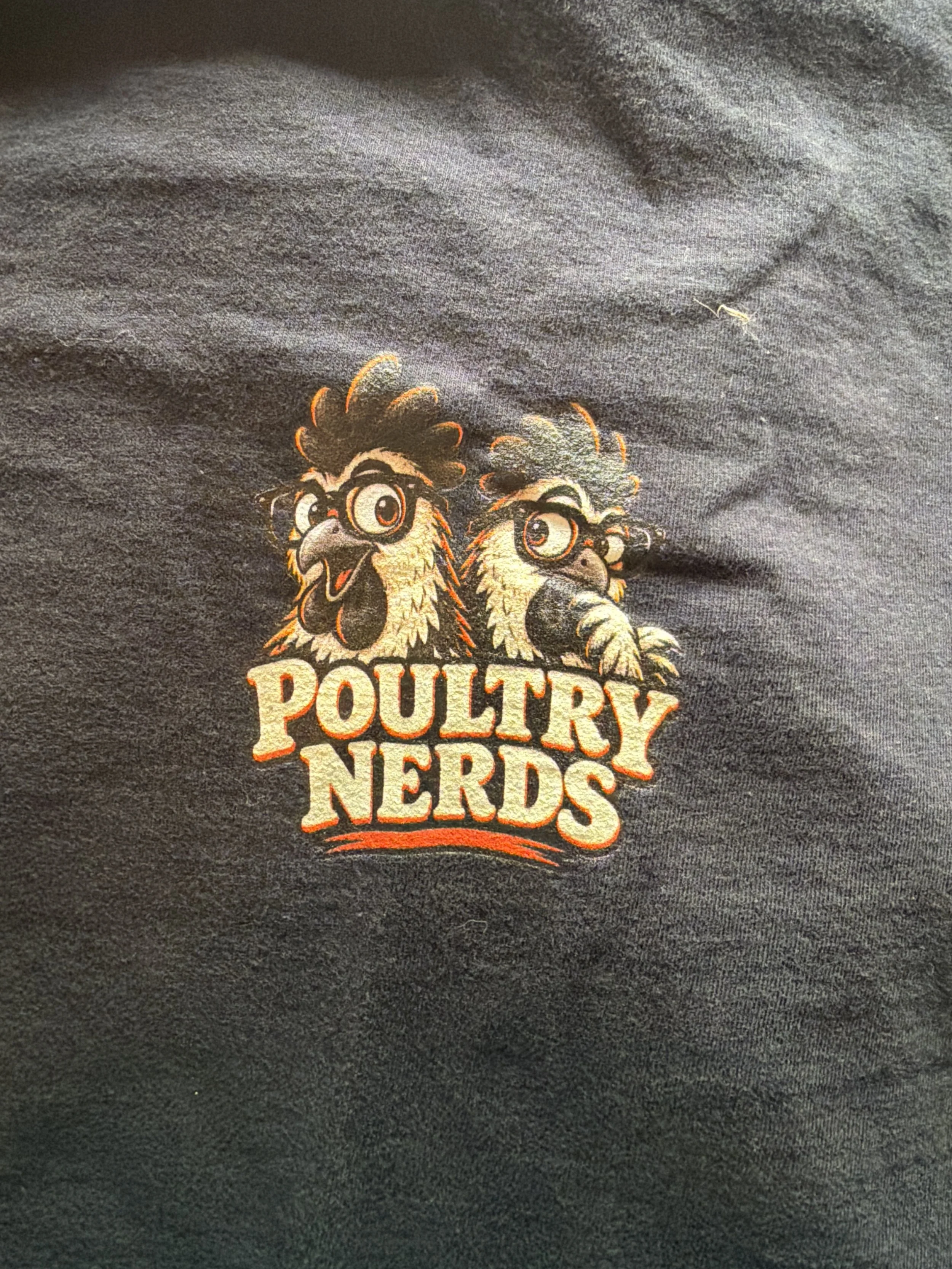Real shirt close-up of the small Poultry Nerds front logo, with two illustrated chickens and cream-and-orange lettering.