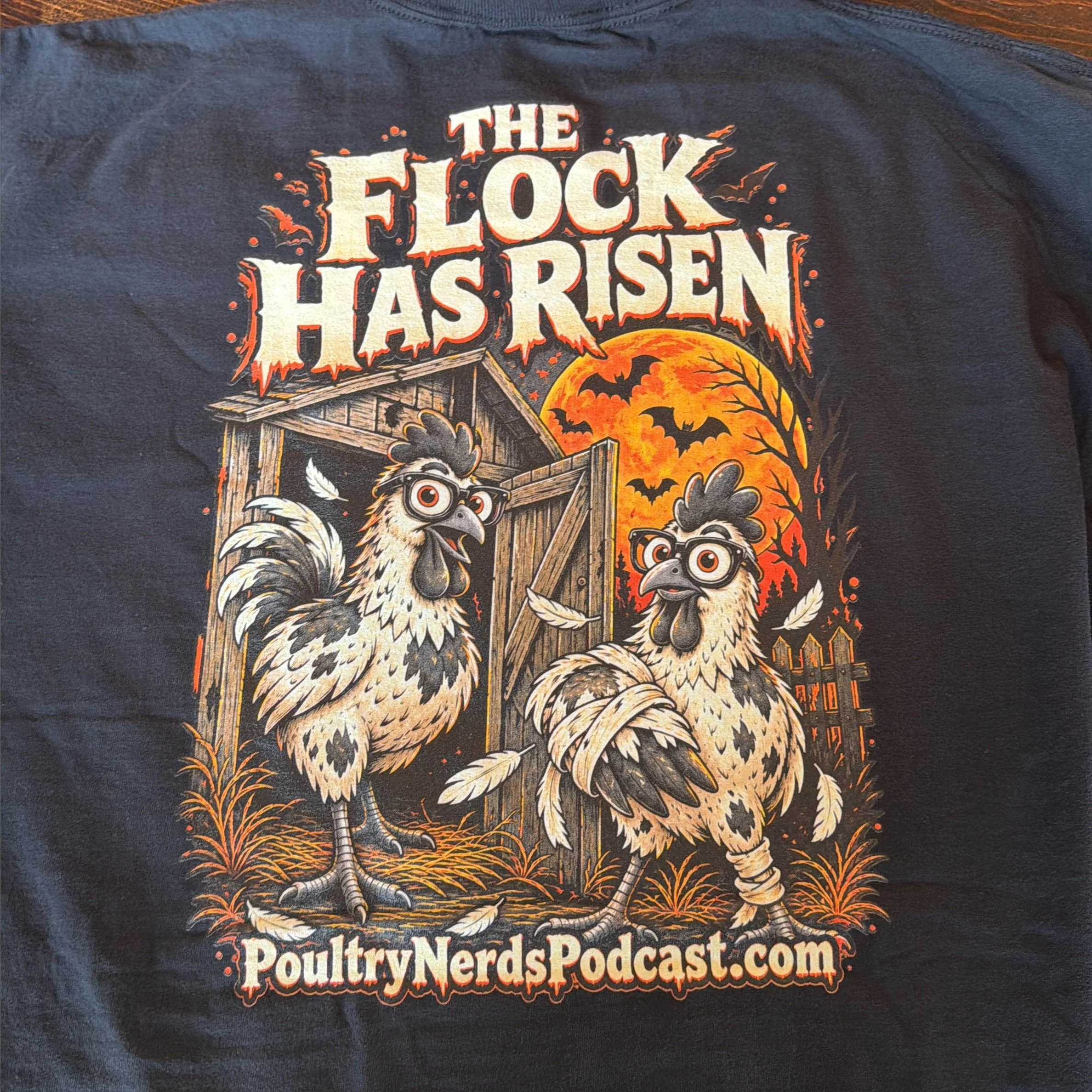Real shirt close-up showing The Flock Has Risen back print with two chickens, an orange moon, bats, and PoultryNerdsPodcast.com.