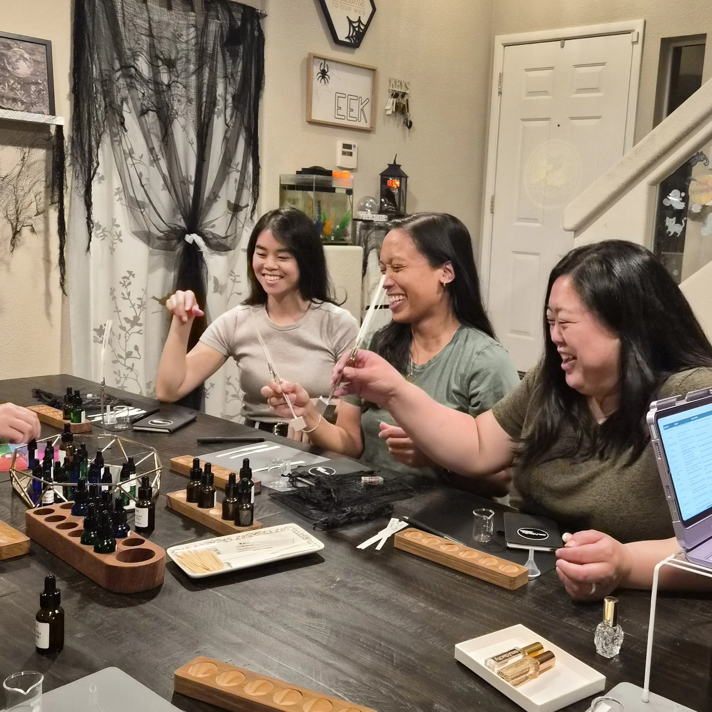 Three women happily engaged in a aromatherapy or essential oil workshop around a black table with various bottles and tools, smiling and laughing. perfume DIY