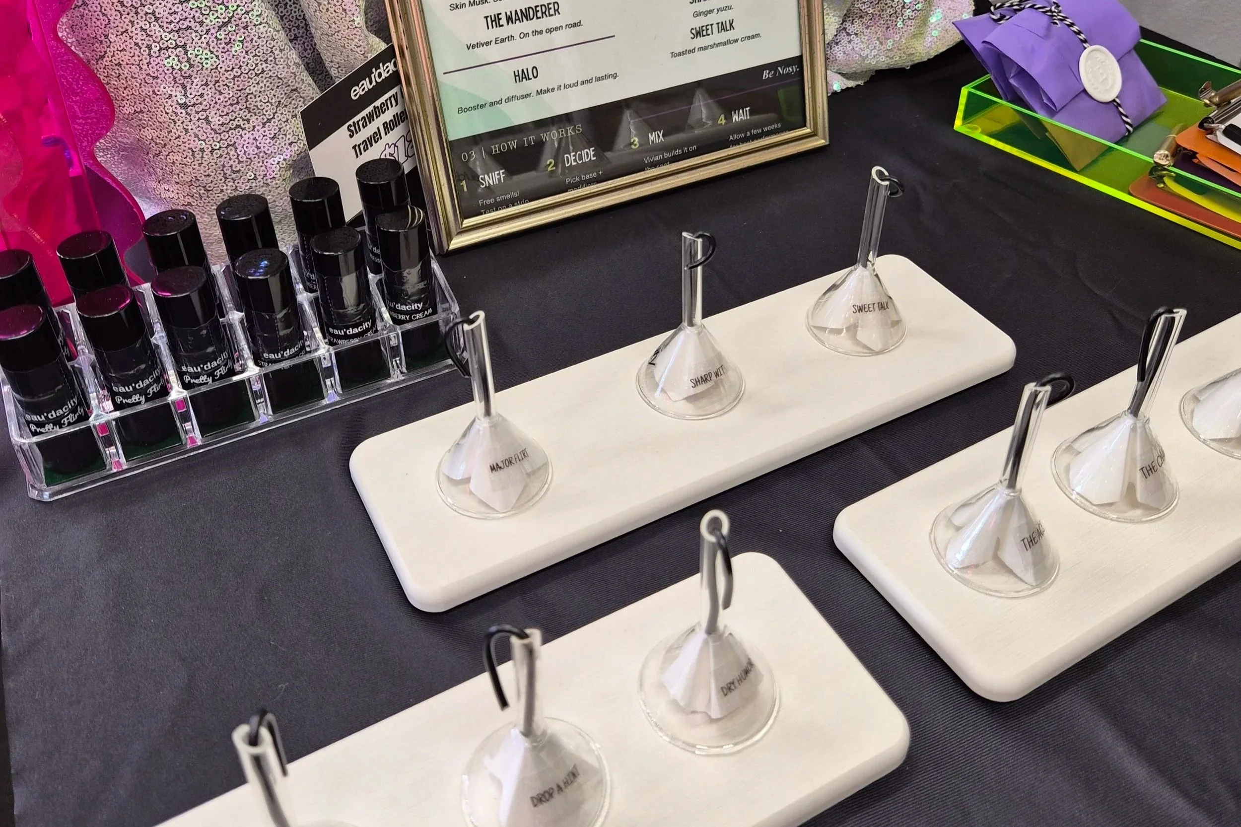 Display of perfume or essential oil samples with black caps in a clear plastic holder, alongside small glass test vials labeled with descriptive words like 'Major Key,' 'Sharp Mint,' 'Drop a Hint,' and 'The One,' on a black table. In the background, there is a framed description with instructions on different scent phases, and colorful items including sequined fabric, a purple wrapped gift, and a green and yellow container with assorted small objects.
