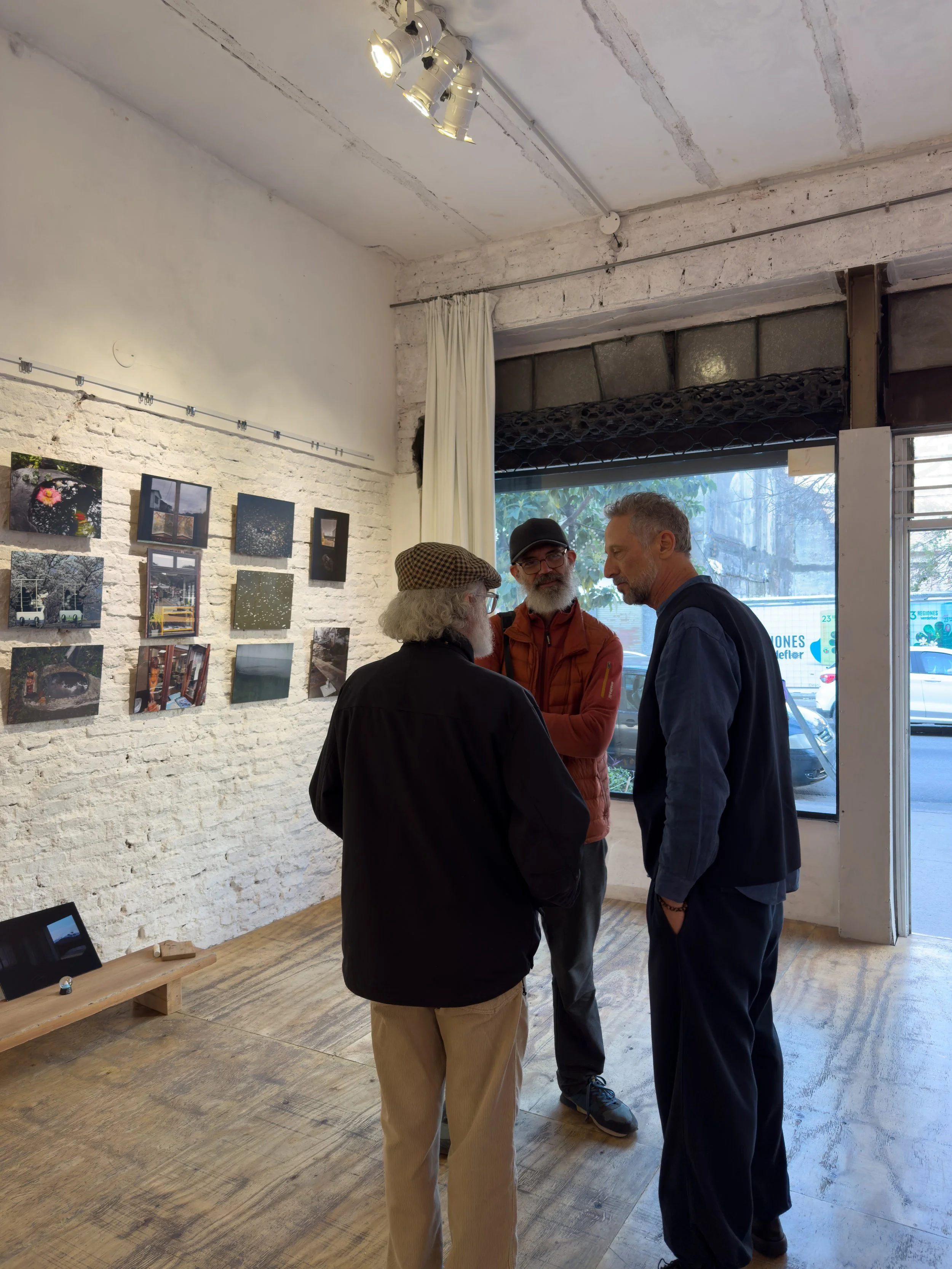 Tres personas conversando en una galería de arte con fotos en la pared, interior con paredes de piedra, suelo de madera y ventana grande.