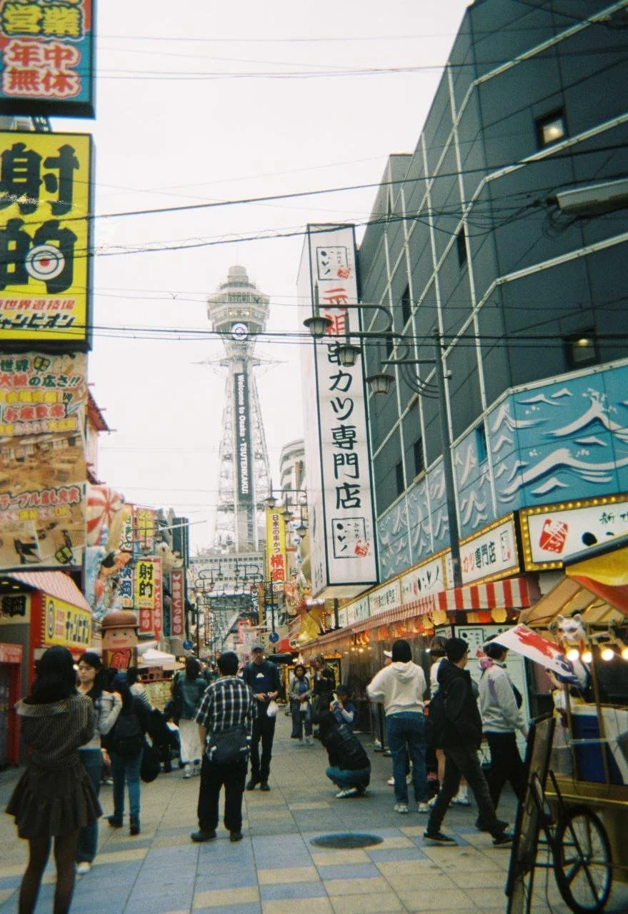 Una calle concurrida en Japón con muchas señalizaciones en japonés, puestos de comida y personas paseando, con la Torre de Tsutenkaku visible en el fondo.