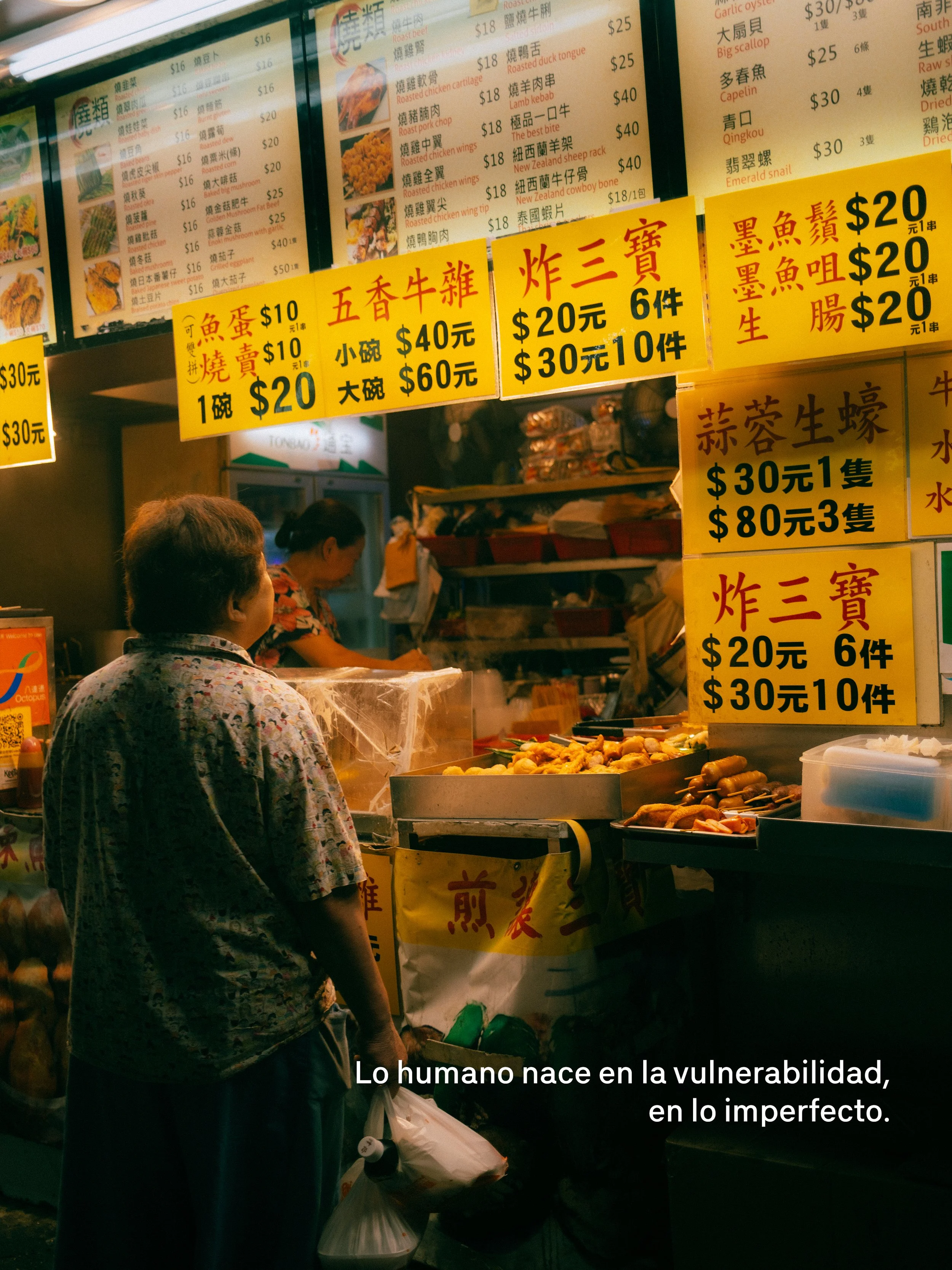 Persona comprando en un puesto callejero de comida, con carteles con precios en chino y algunos en español, que venden diferentes tipos de alimentos y mariscos en un entorno nocturno.