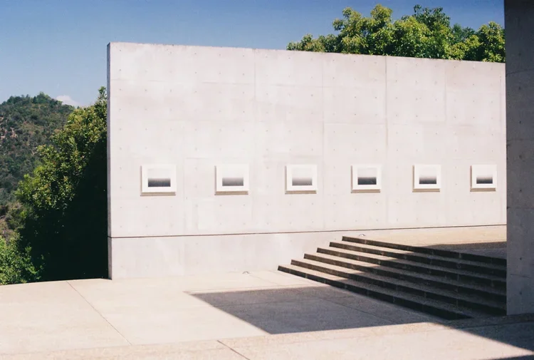 Edificio moderno de concreto con ventanas pequeñas en una pared blanca, escaleras de piedra y árboles verdes al fondo.
