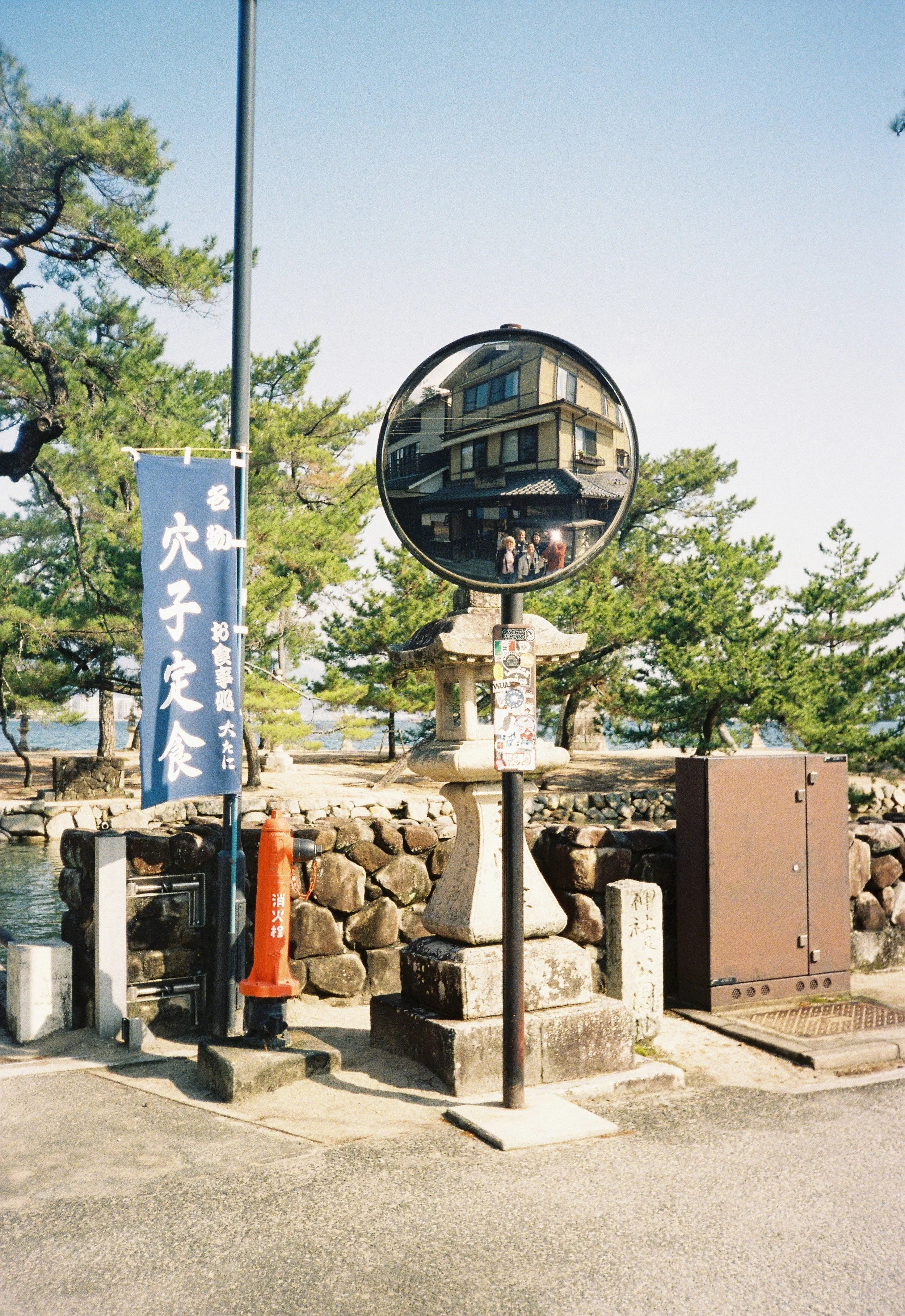 Espejo de tráfico que refleja un edificio tradicional japonés, árboles y algunas personas en un parque junto a un cuerpo de agua, con un cielo despejado y algunos carteles y objetos en la zona.