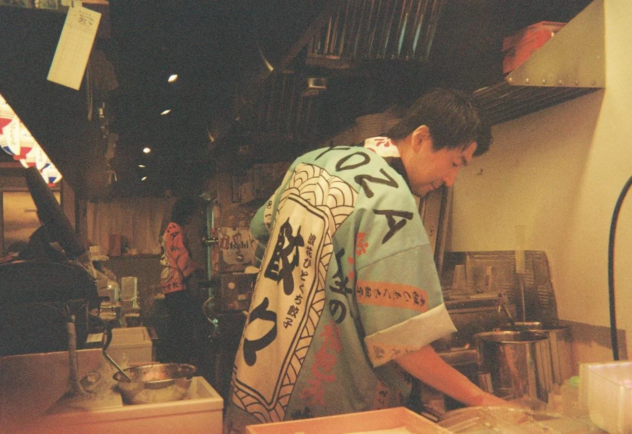 Hombre preparando comida en una cocina de restaurante japonés, usando un delantal tradicional. Hay utensilios y ingredientes en la mesa y estanterías con artículos en el fondo.
