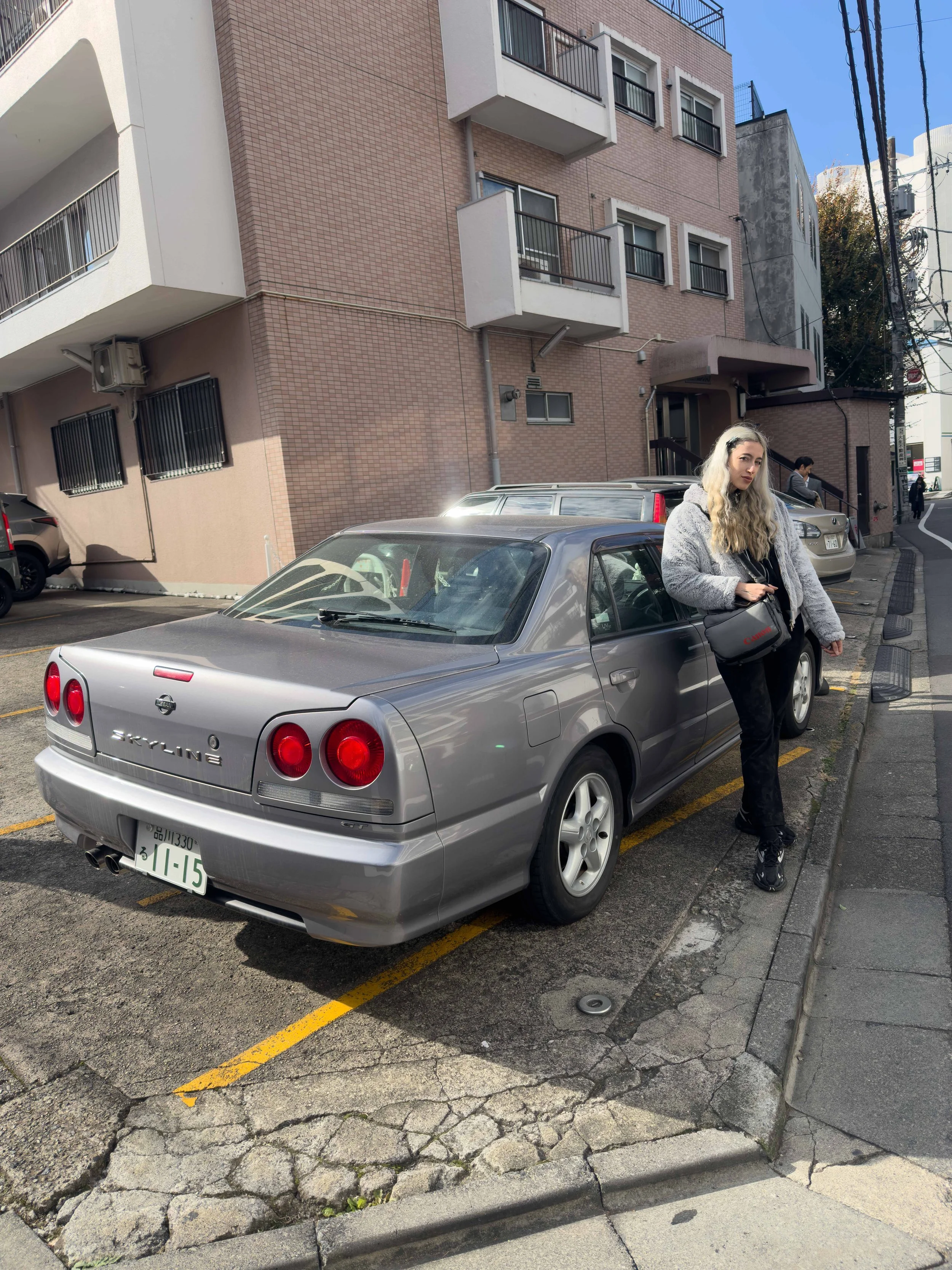 Una joven con cabello rubio y ondulado, vestida con sudadera gris y pantalones negros, está de pie junto a un coche plateado en una calle urbana con edificios residenciales y varias personas de fondo.