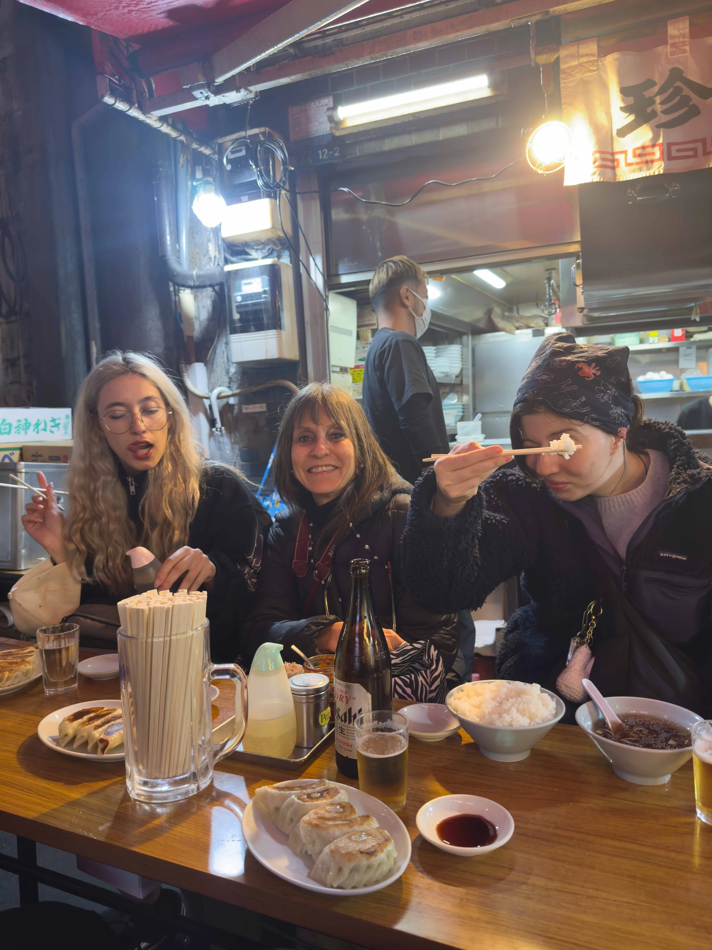 Personas comiendo en un restaurante de comida japonesa, hay sushi, ramen, arroz, yotros platillos en la mesa, paredes y zona de cocina en el fondo, ambiente informal.