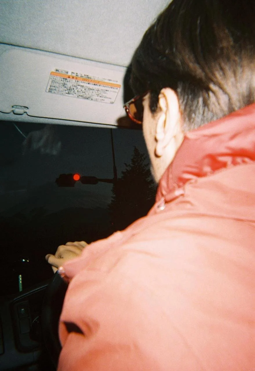 Persona conduciendo un automóvil por la noche, vista de espaldas, con una gorra, gafas de sol y chaqueta roja.