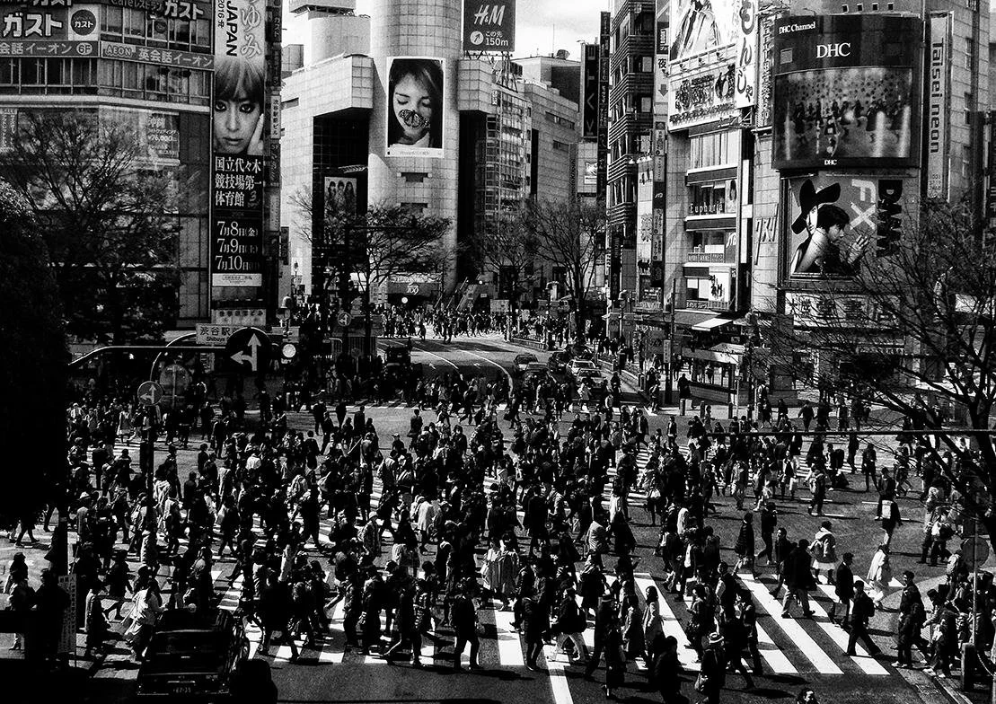 Una multitud de personas cruzando en un cruce peatonal en una zona urbana con edificios altos y anuncios publicitarios en japonés.