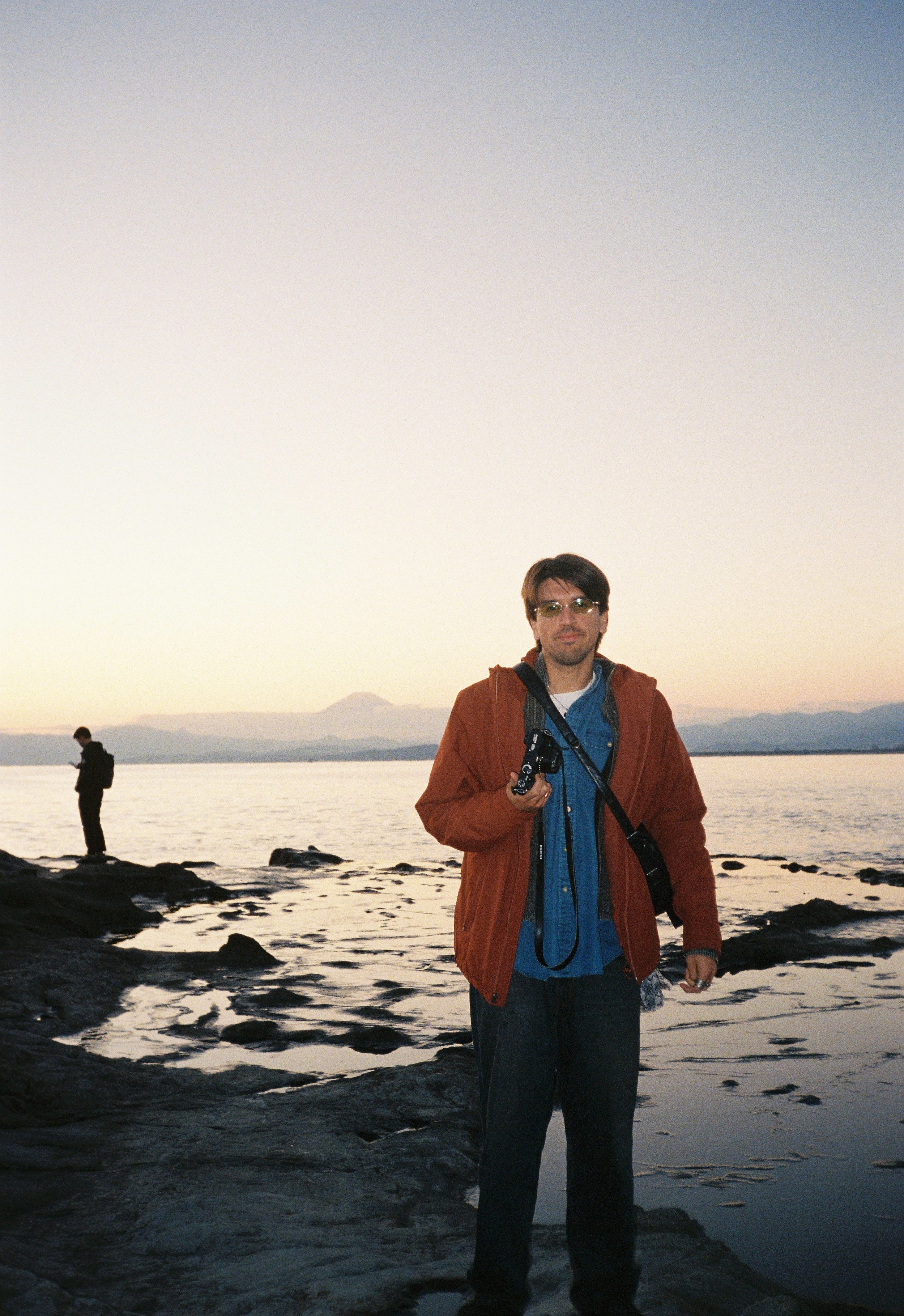 Hombre con gafas y chaqueta anaranjada de pie junto al agua en un paisaje al atardecer, con dos personas más en el fondo cerca de las rocas.