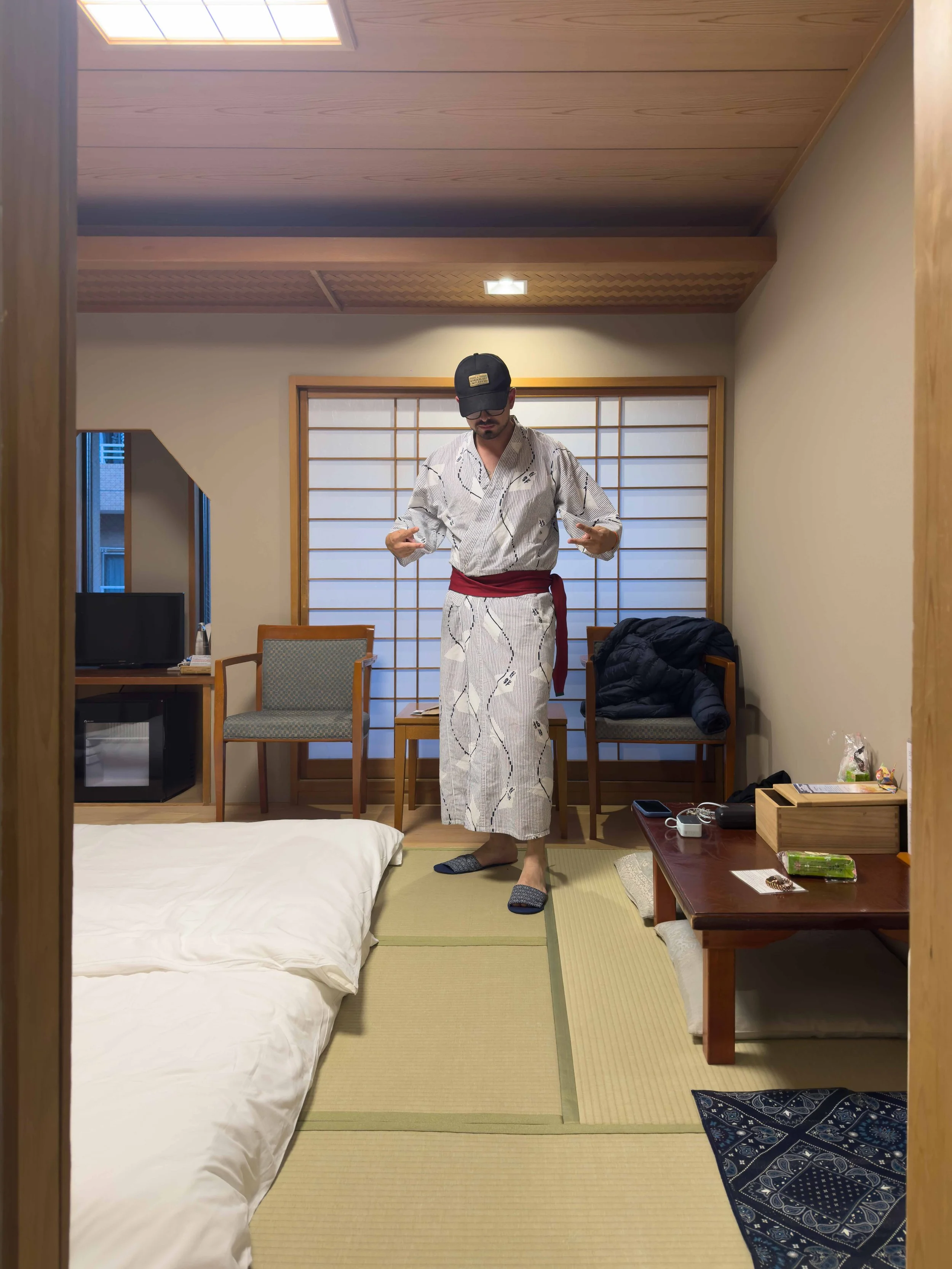 Hombre con vestido tradicional japonés y gorra, en una habitación con puertas correderas de papel, sentado y ajustándose su ropa.