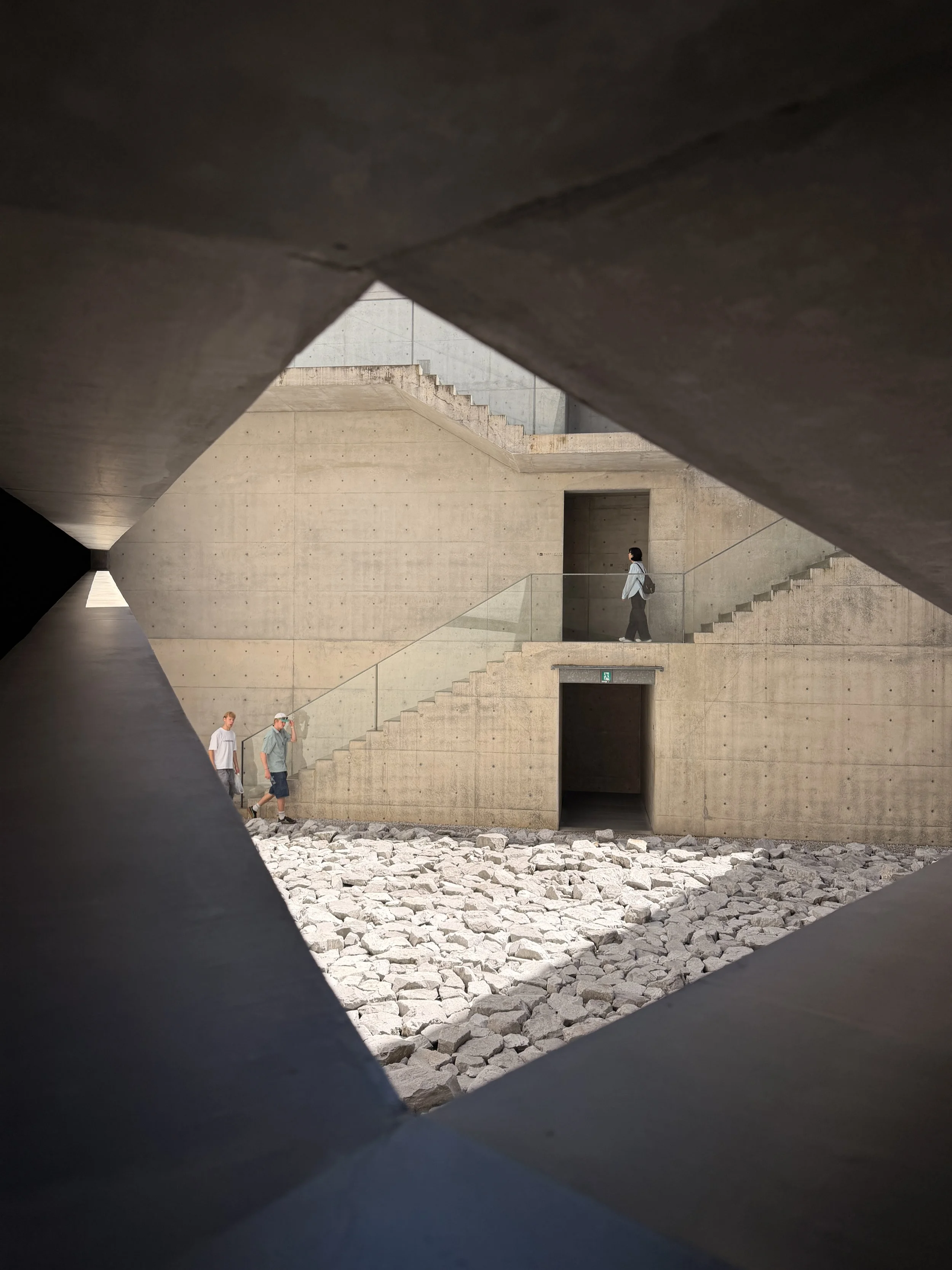 Imagen de una estructura moderna de concreto con escaleras y una pared, vista a través de un marco angular, con tres personas caminando en el espacio.