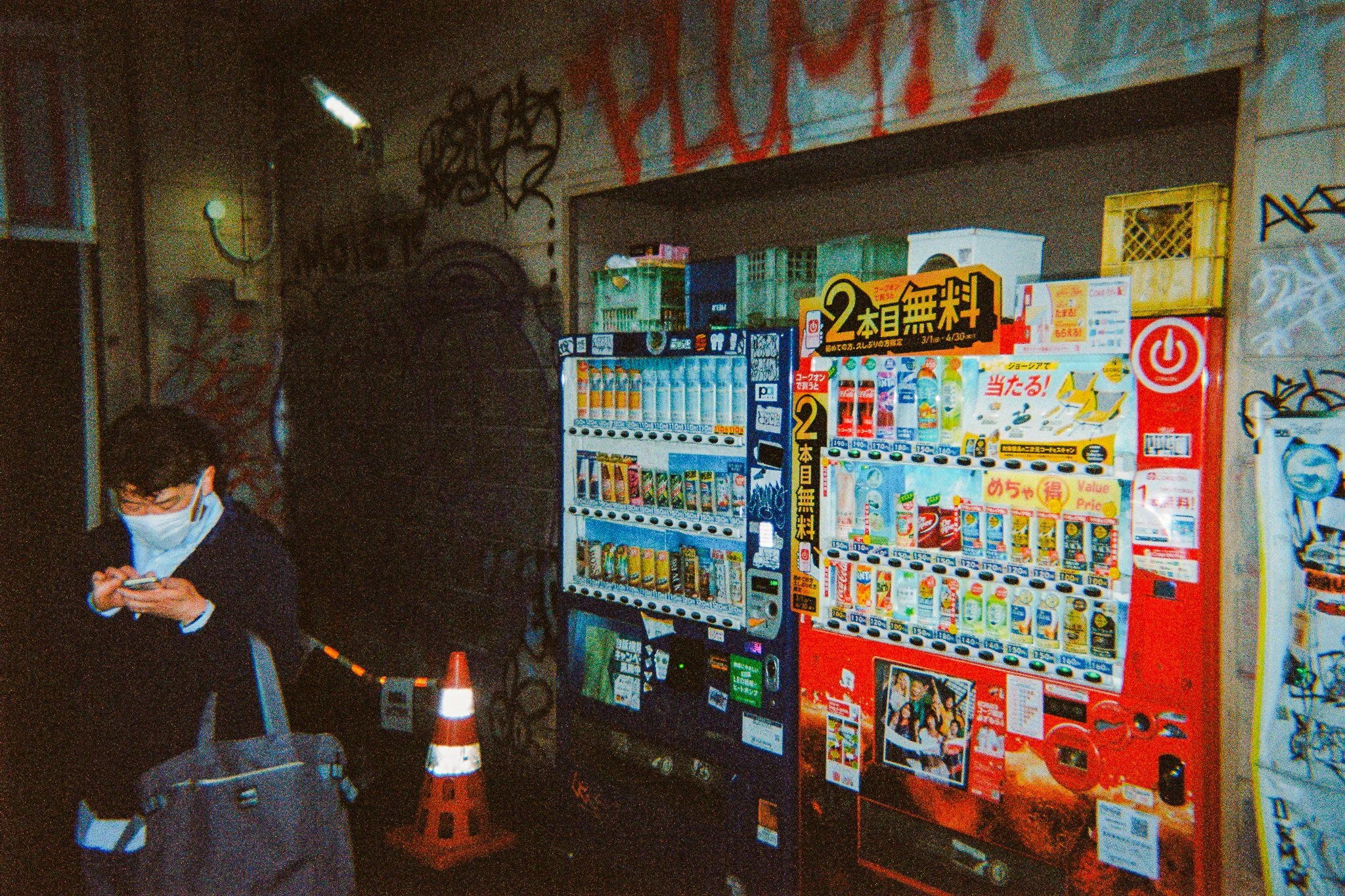 Una mujer con mascarilla revisando su teléfono en una tienda o estación de servicio con máquinas expendedoras y cartelera con información y productos en japonés.