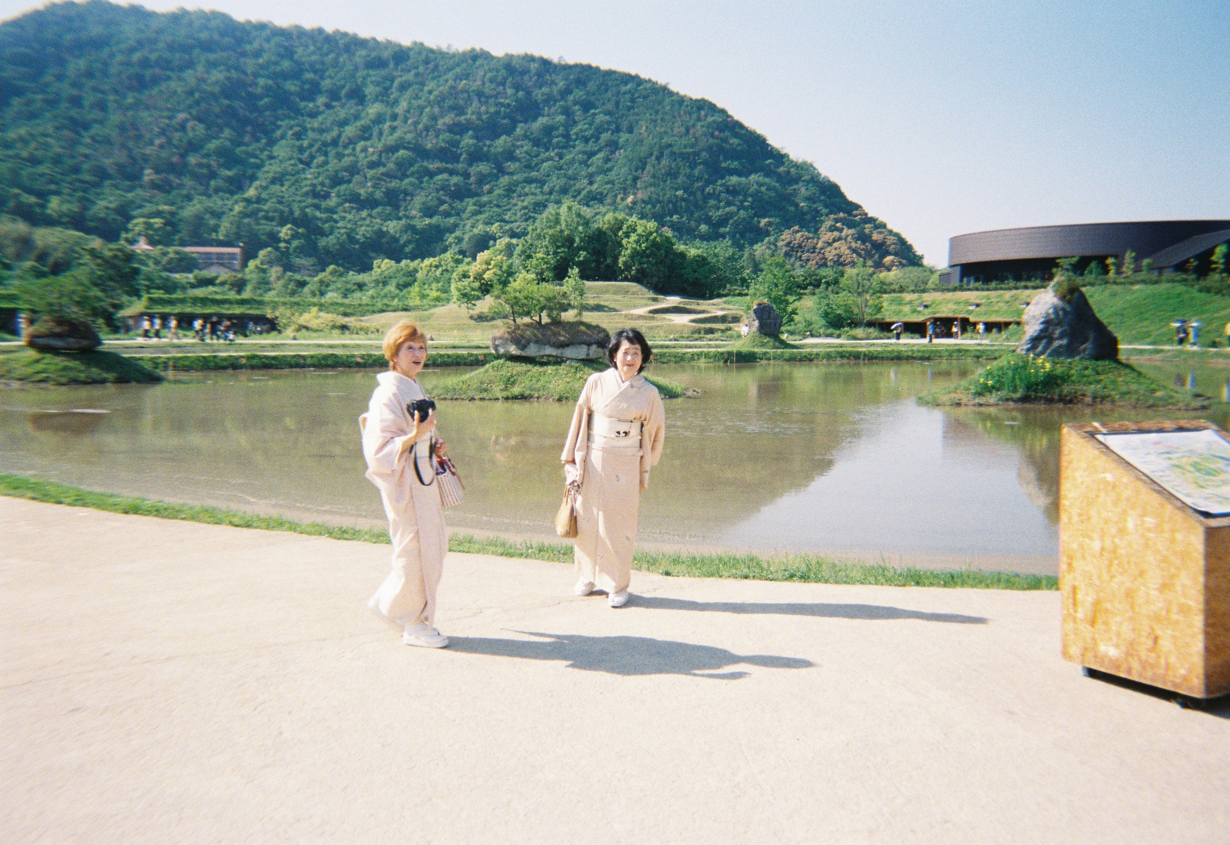 Dos mujeres vestidas con kimono japonés caminando cerca de un lago, con un paisaje montañoso y un edificio moderno en el fondo en un día soleado.