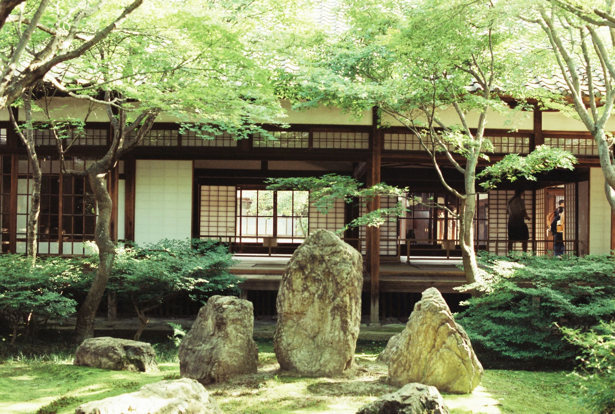 Vista de un jardín con árboles verdes y cabaña tradicional japonesa de madera y puertas correderas de papel.