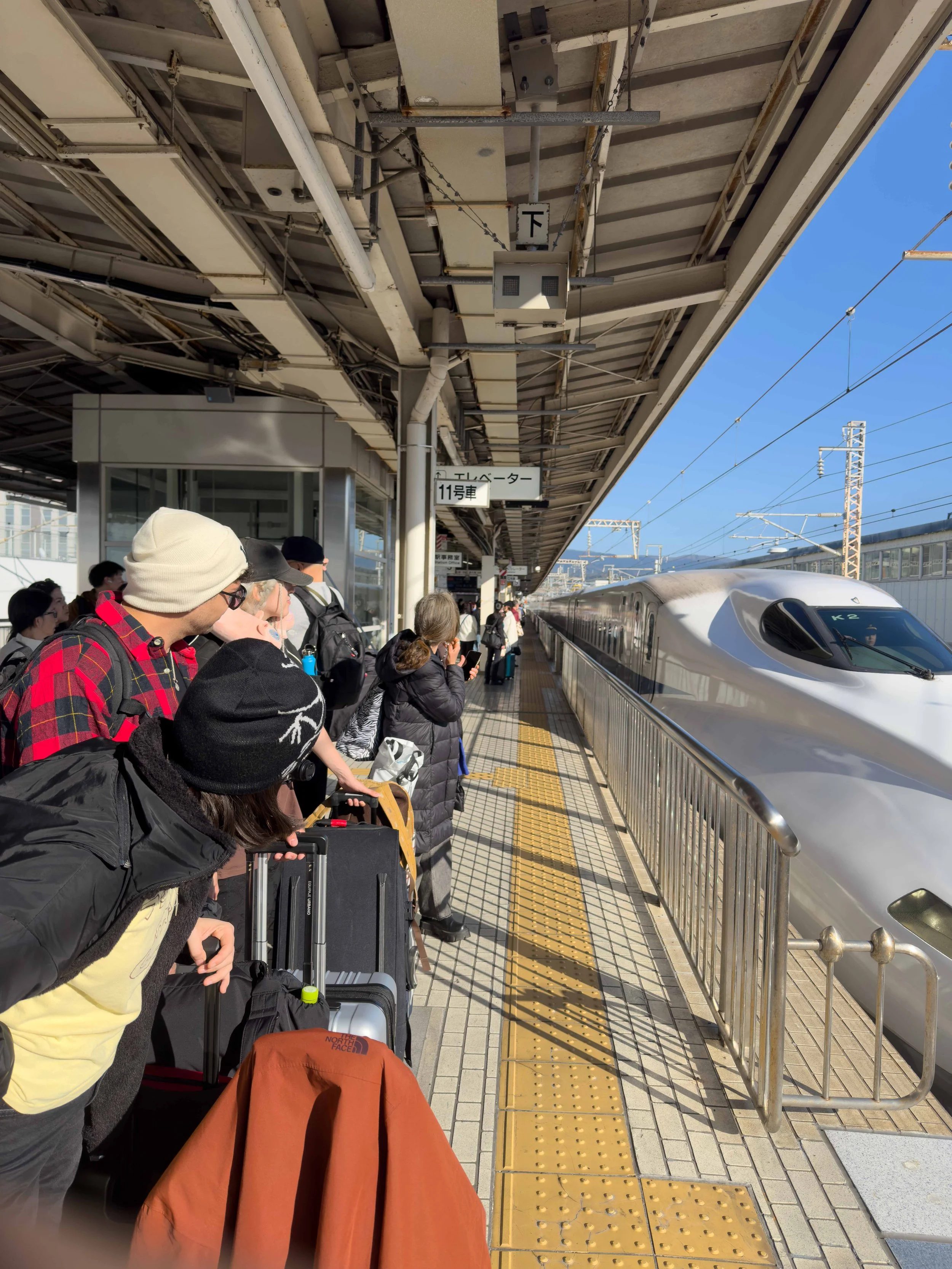Pasajeros en la estación de tren esperando abordar el tren de alta velocidad, con señalización en japonés y gente con equipaje.
