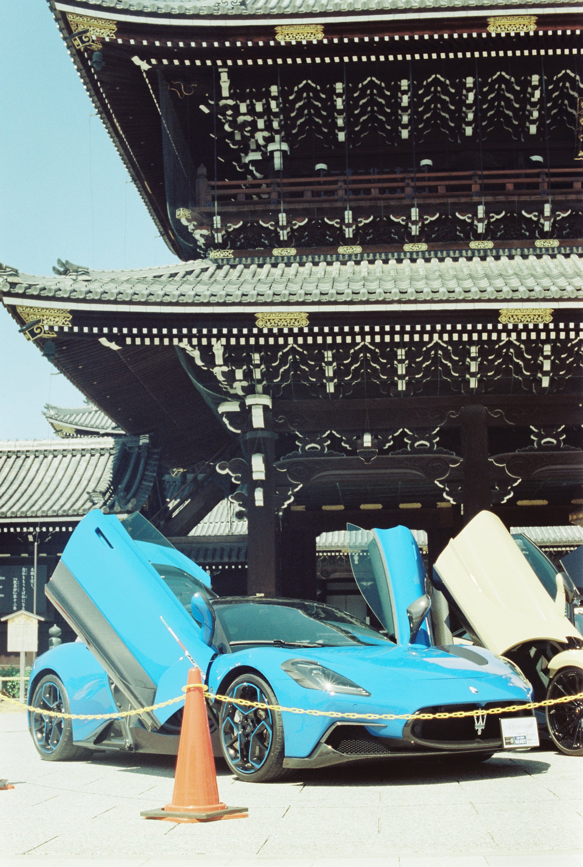 Coche deportivo azul con las puertas abiertas en exhibición en un evento al aire libre. Frente al coche hay un cono de tráfico naranja y una cadena amarilla delimitando el área.