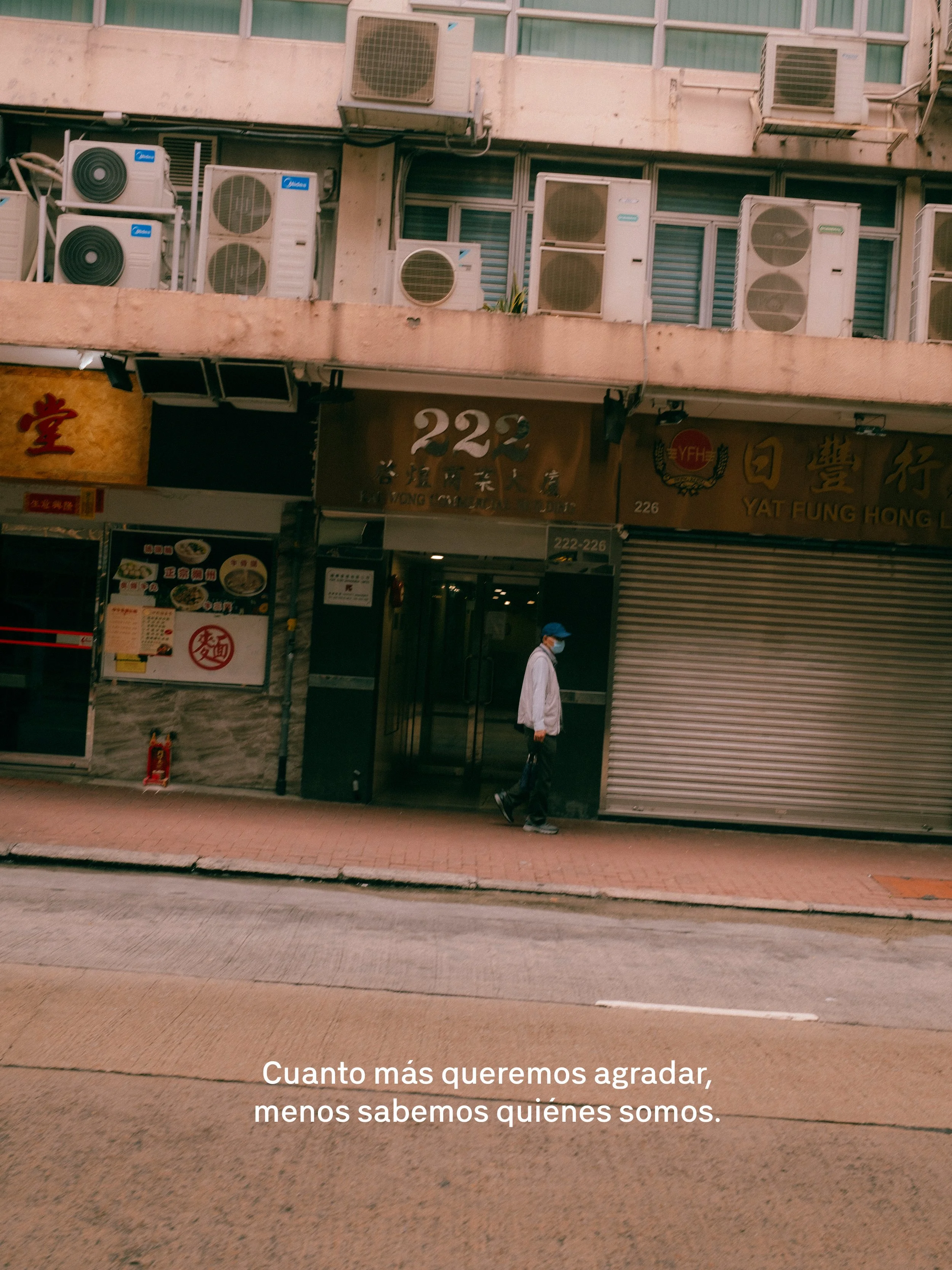 Foto de un edificio con tiendas en la planta baja y varias unidades de aire acondicionado en el segundo piso, con un hombre usando mascarilla caminando por la acera. En la parte inferior, texto que dice: "Cuanto más queremos agradar, menos sabemos quiénes somos."
