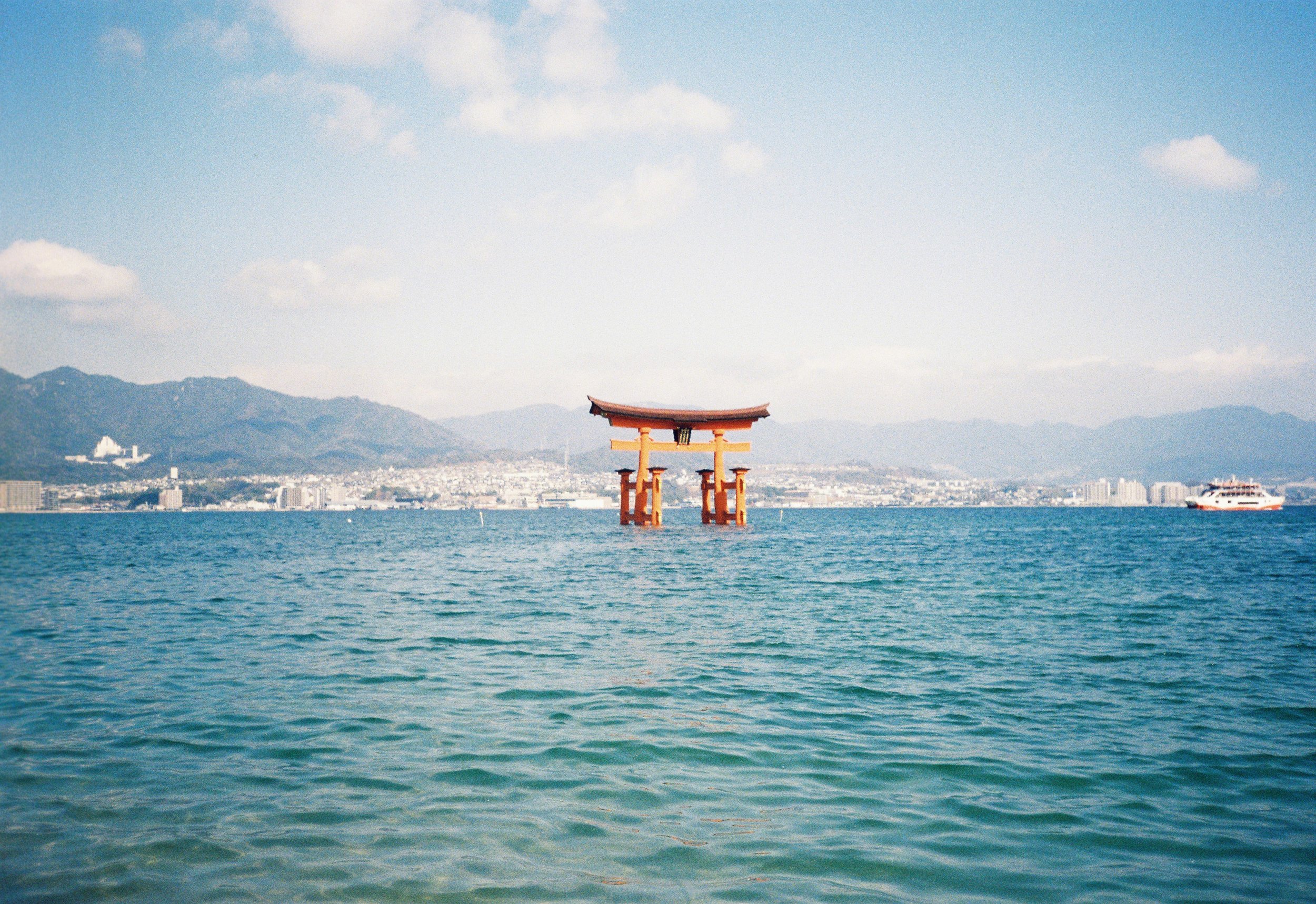 Una vista del mar con un torii japonés en el agua y una ciudad de fondo con montañas y edificios.