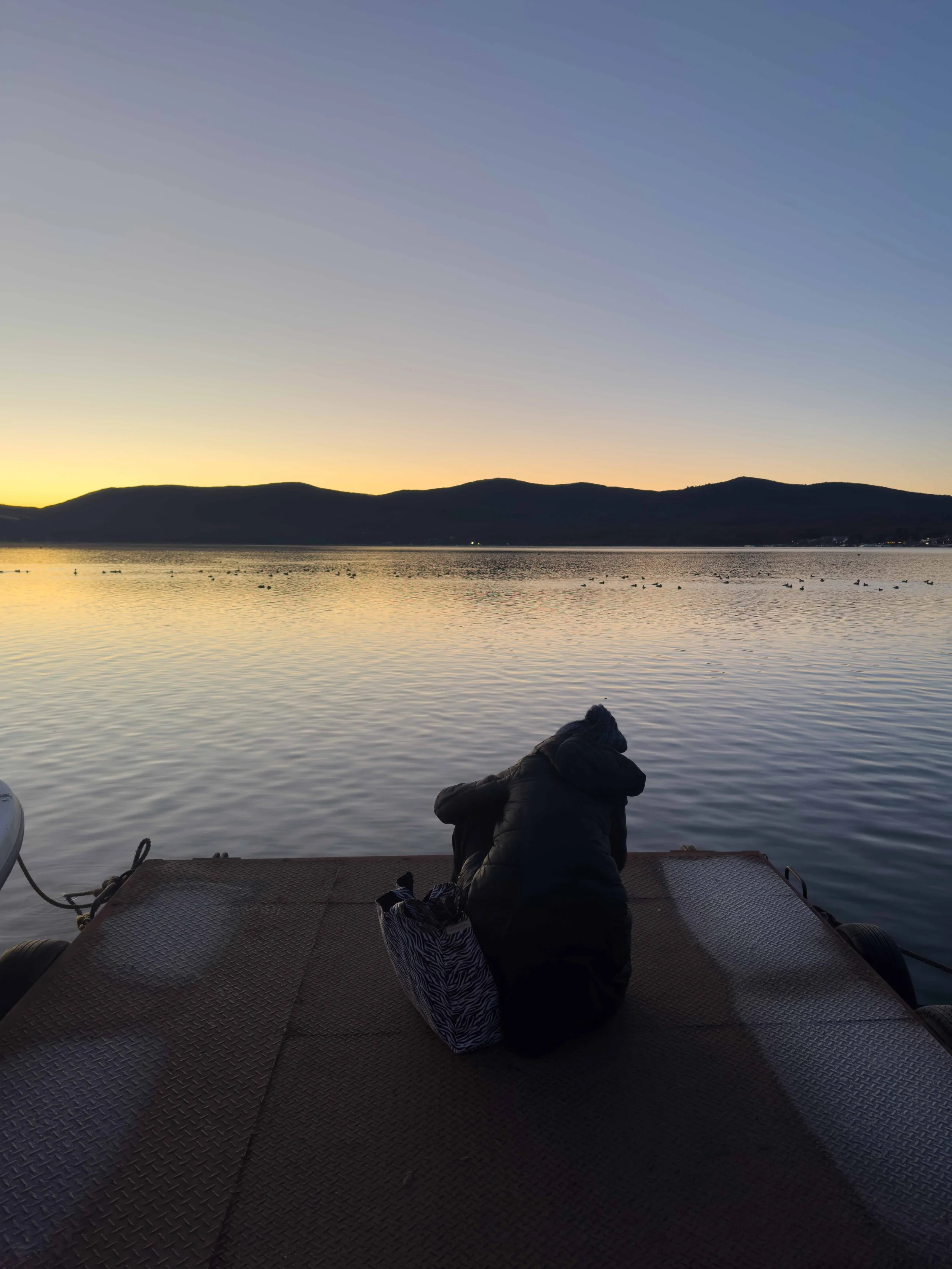 Persona agachada en una plataforma junto a un cuerpo de agua al atardecer, con montañas y un cielo claro en el fondo.