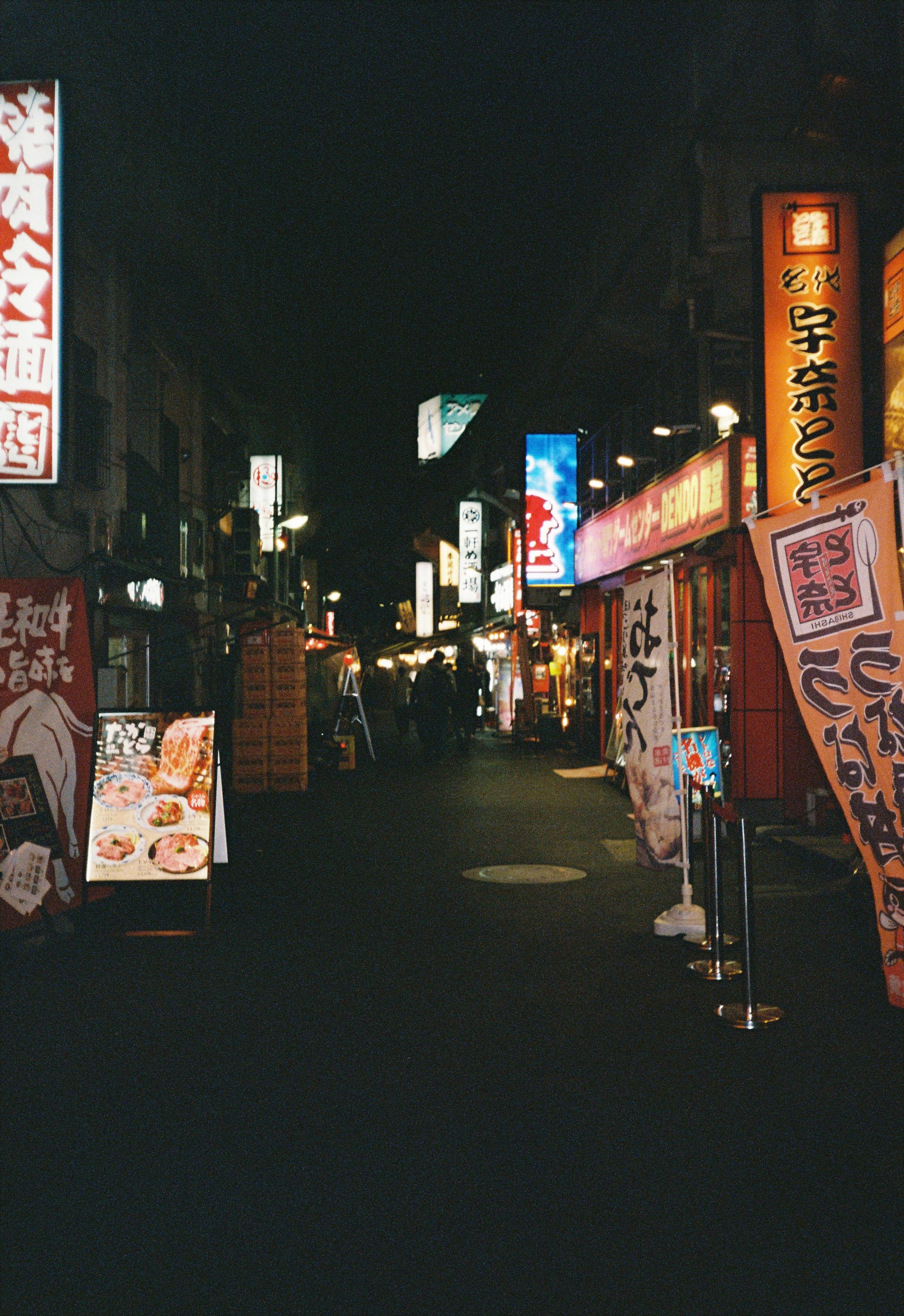 Una calle peatonal en Japón con letreros luminosos en japonés, tiendas y restaurantes, y carteles con imágenes de comida en la noche.