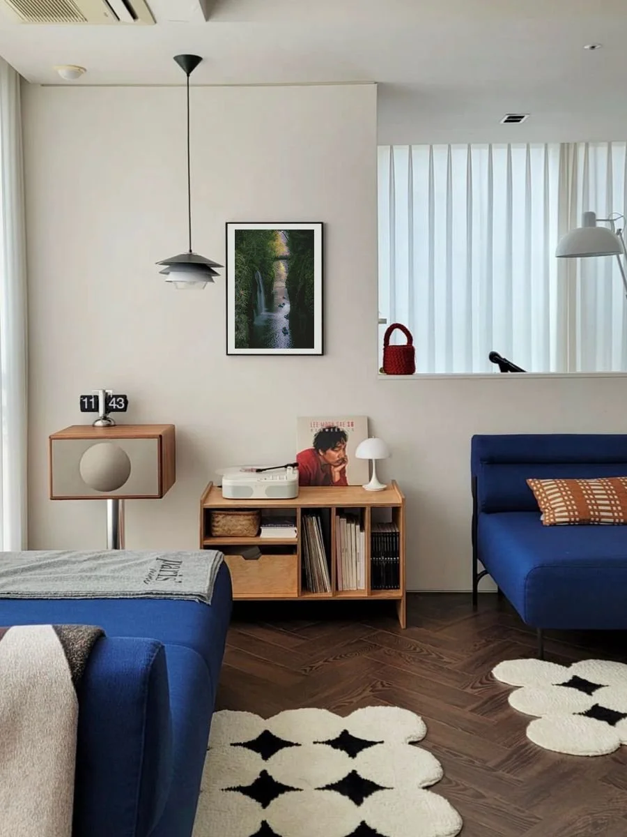 Sala de estar con paredes blancas, una lámpara de techo negra, un cuadro de naturaleza, una estantería con discos, libros y decoraciones, un sofá azul, una alfombra blanca con motivos negros y una ventana con cortinas blancas.