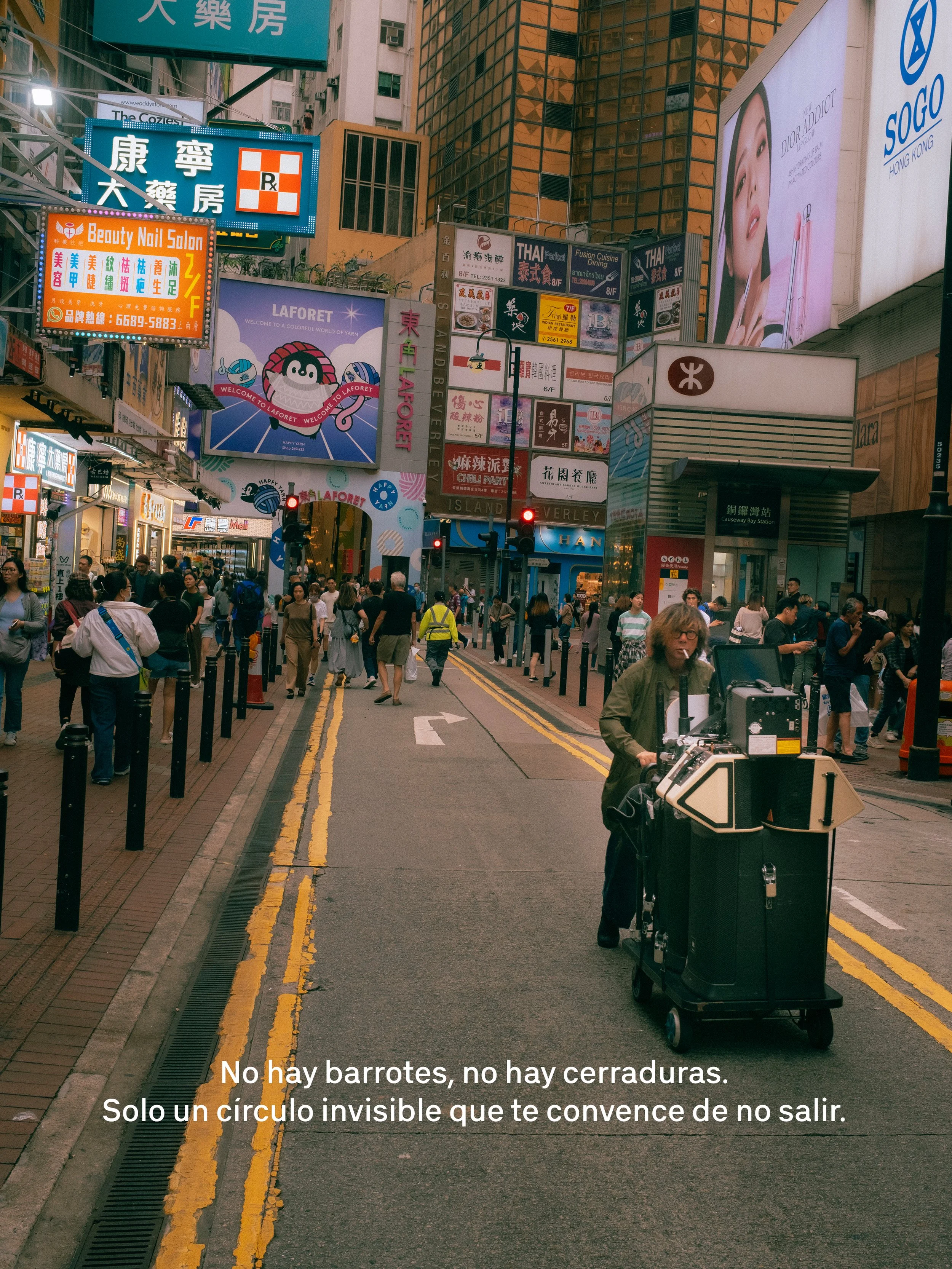 Una bulliciosa calle en Hong Kong con muchas señales y anuncios de neón, y personas caminando en la acera. En primer plano, una persona con una máquina de rebanar comida y una maleta, y un carril con marcas en el suelo.