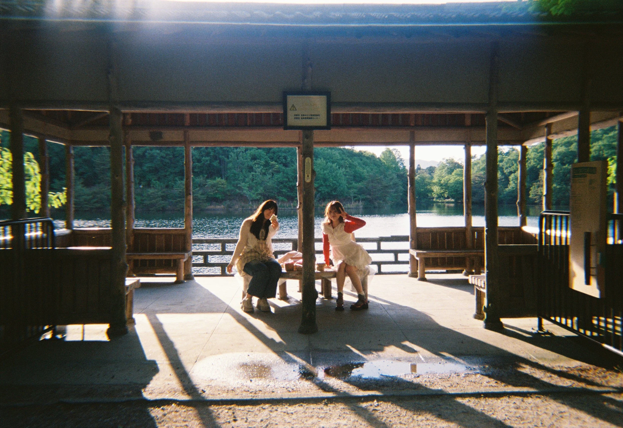Dos niñas sentadas en un banco de madera bajo un toldo junto a un río rodeado de árboles verdes en un día soleado.