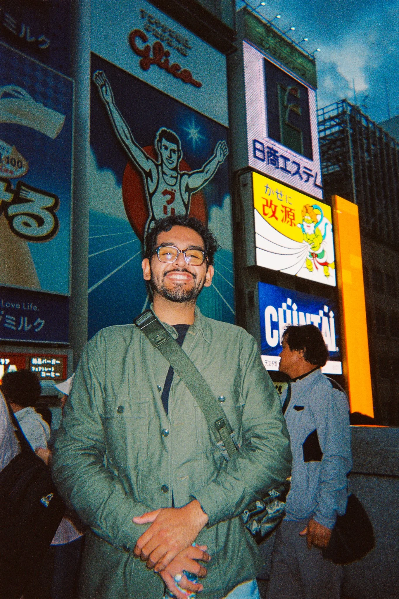 Un hombre con gafas y chaqueta verde en una ciudad asiática, con carteles y publicidad en letras japonesas y anuncios en la noche.