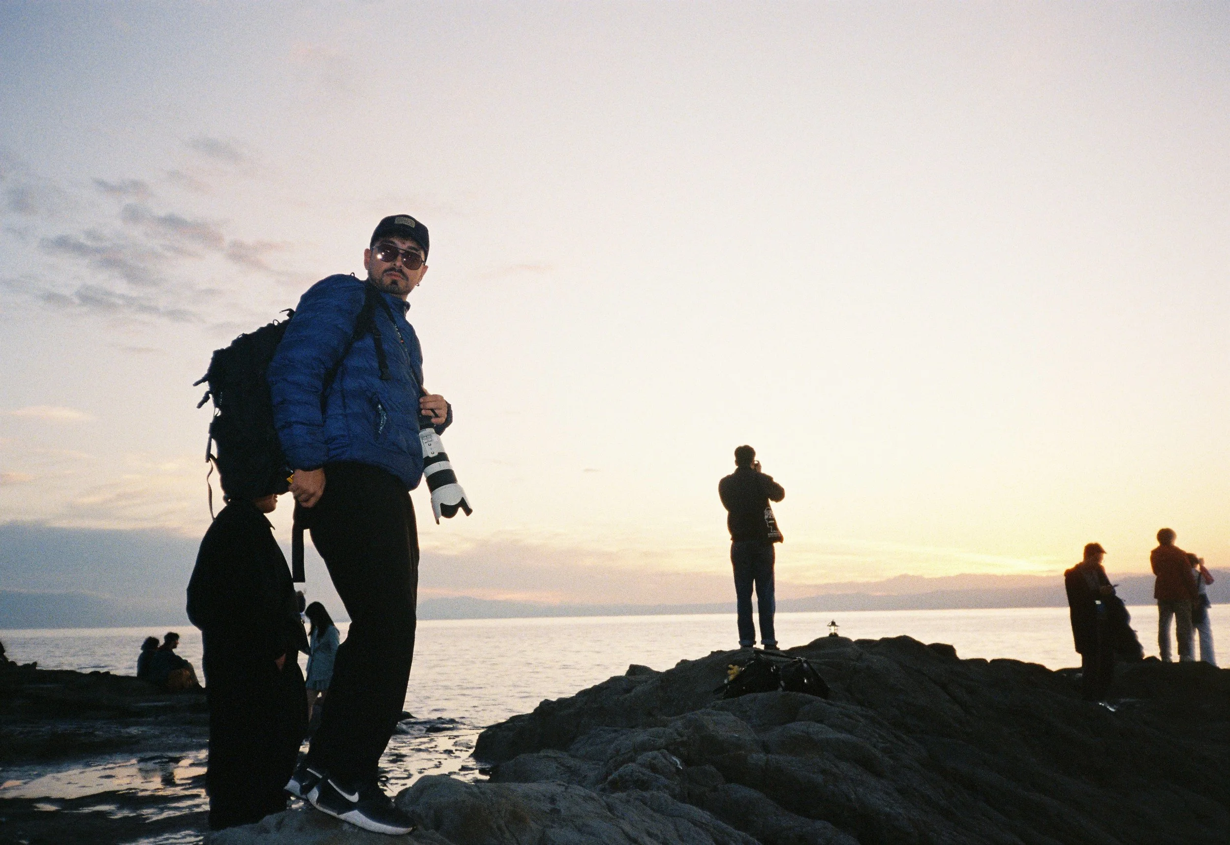 Un hombre con gafas de sol, chaqueta azul y mochila de senderismo de pie en la orilla del mar al atardecer, con otras personas en el fondo disfrutando del paisaje.